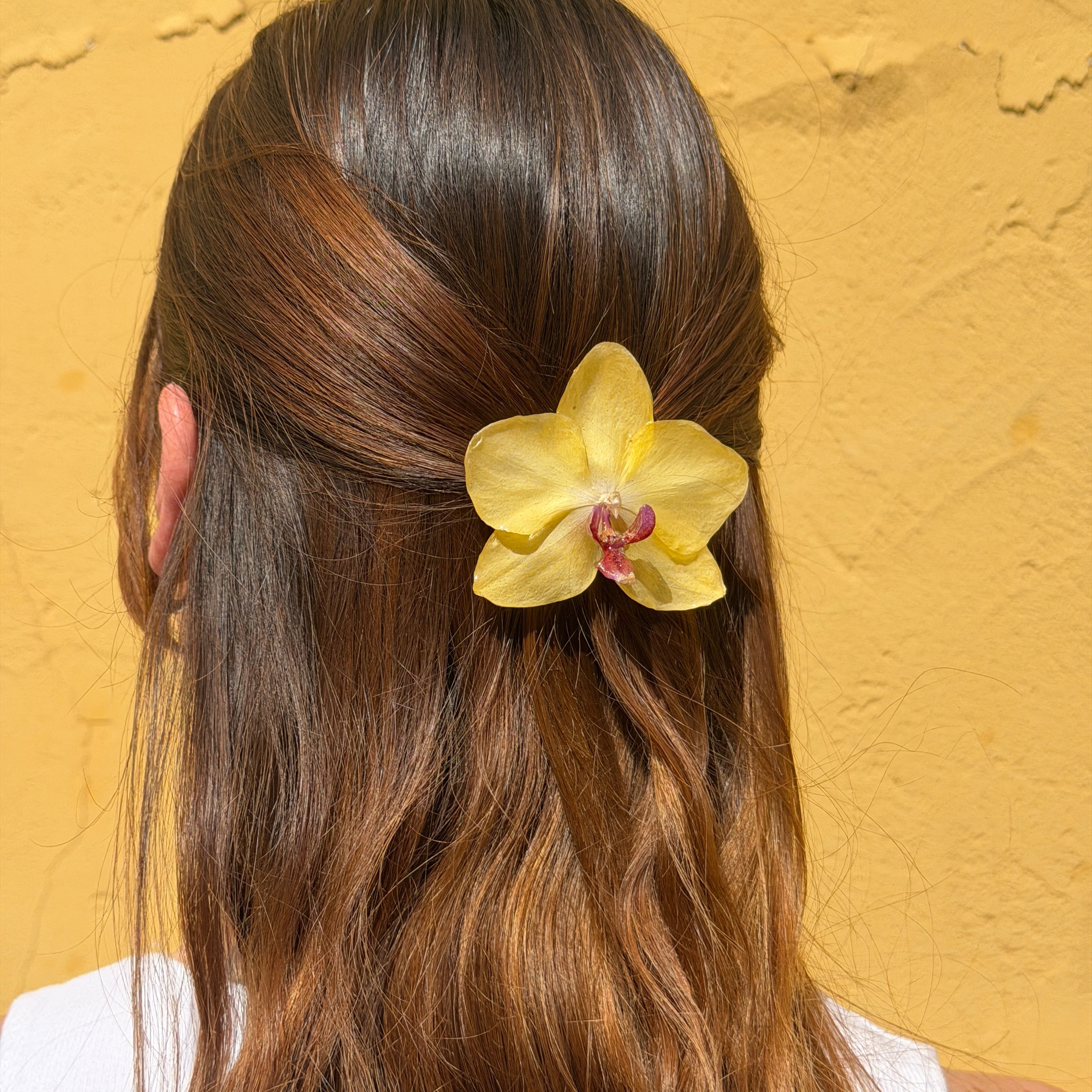 Pinza de pelo con orquídea amarilla preservada en resina colocada sobre el cabello castaño de una mujer, con fondo amarillo cálido.