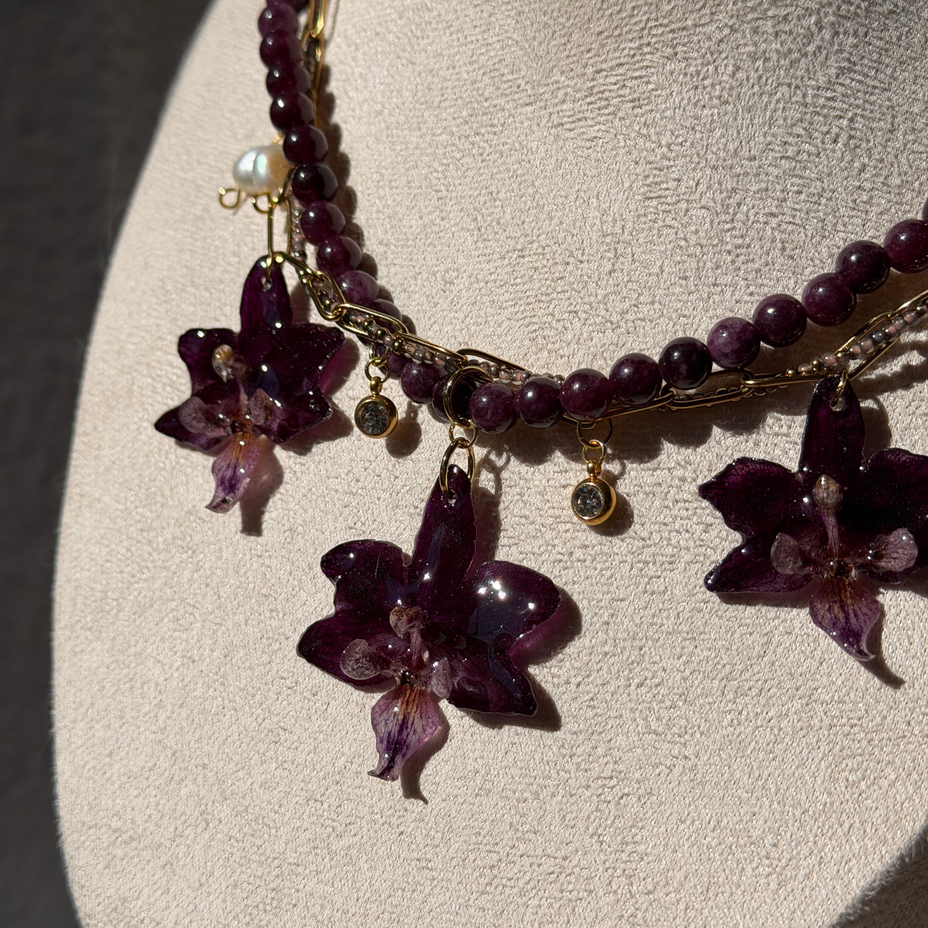 Collar Orquídea Ritual en busto expositor mostrando las tres orquídeas púrpura preservadas en resina con cadena triple dorada, perlas de agua dulce y charms de cristal