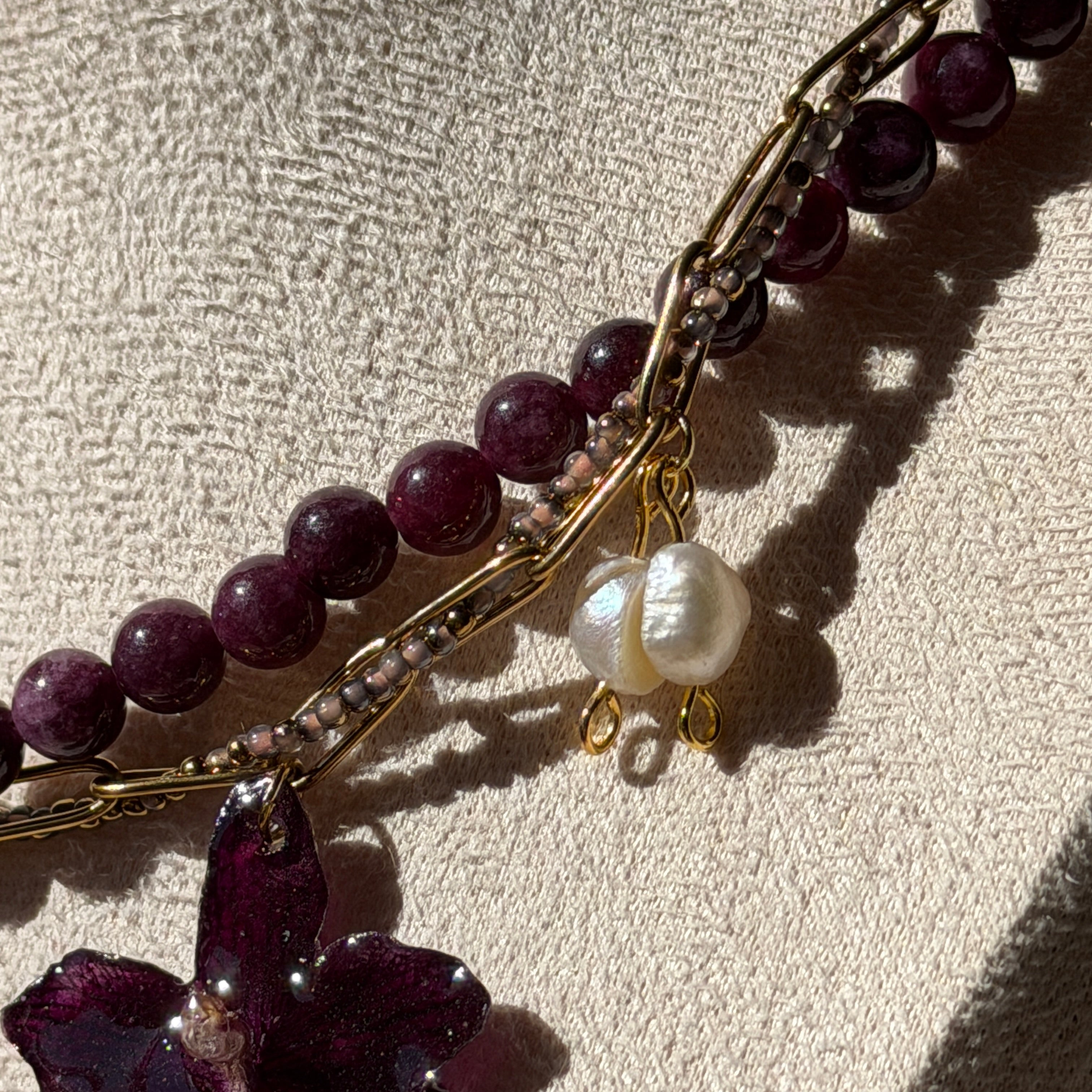 Detalle macro del Collar Orquídea Ritual mostrando las piedras de cristal púrpura, cadena de eslabones dorados, microperlas y perla natural de agua dulce sobre plato cerámico