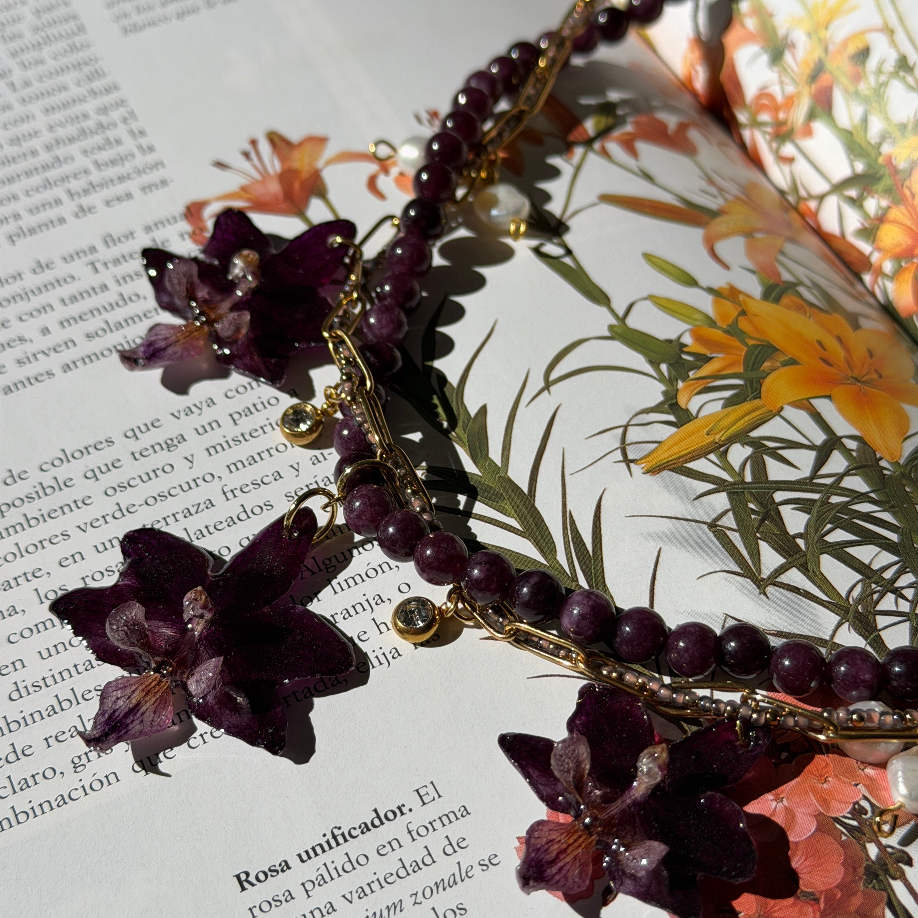 Collar Orquídea Ritual de Aurum Orchid con tres orquídeas púrpura preservadas en resina sobre libro botánico vintage, cadena triple dorada con piedras de cristal y perlas de agua dulce