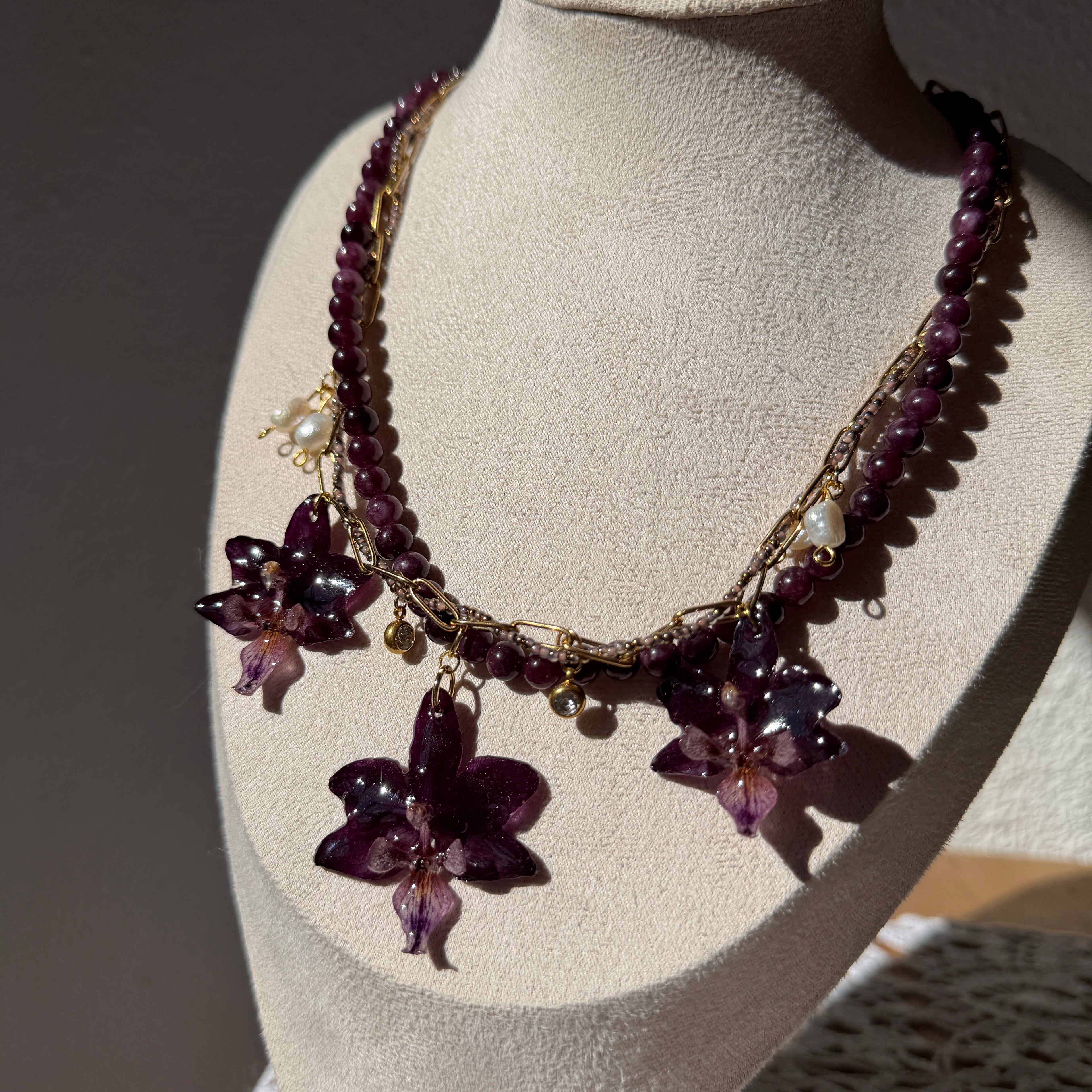 Detalle frontal del Collar Orquídea Ritual en busto expositor con tres orquídeas púrpura preservadas en resina, perla de agua dulce y charms de cristal engarzado dorado