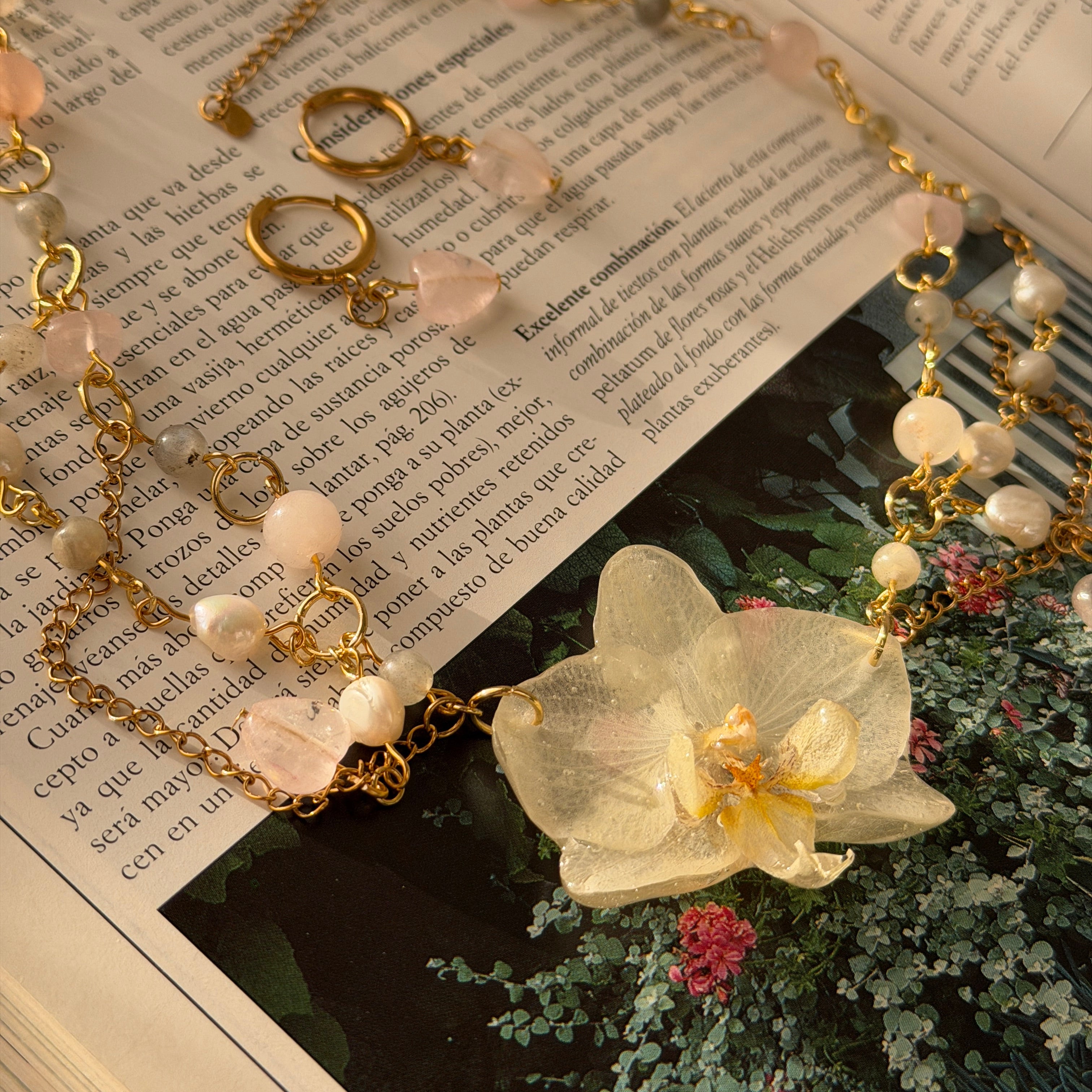 Set Orquídea Estela completo en flat lay sobre libro vintage: collar con orquídea phalaenopsis preservada en resina, cadena de perlas naturales y cuarzo rosa con pendientes huggie dorados
