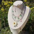Set Margarita Brisa en busto expositor entre flores silvestres amarillas, mostrando la cadena doble dorada con piedras transparentes, flores de acrílico turquesa y margarita preservada en resina