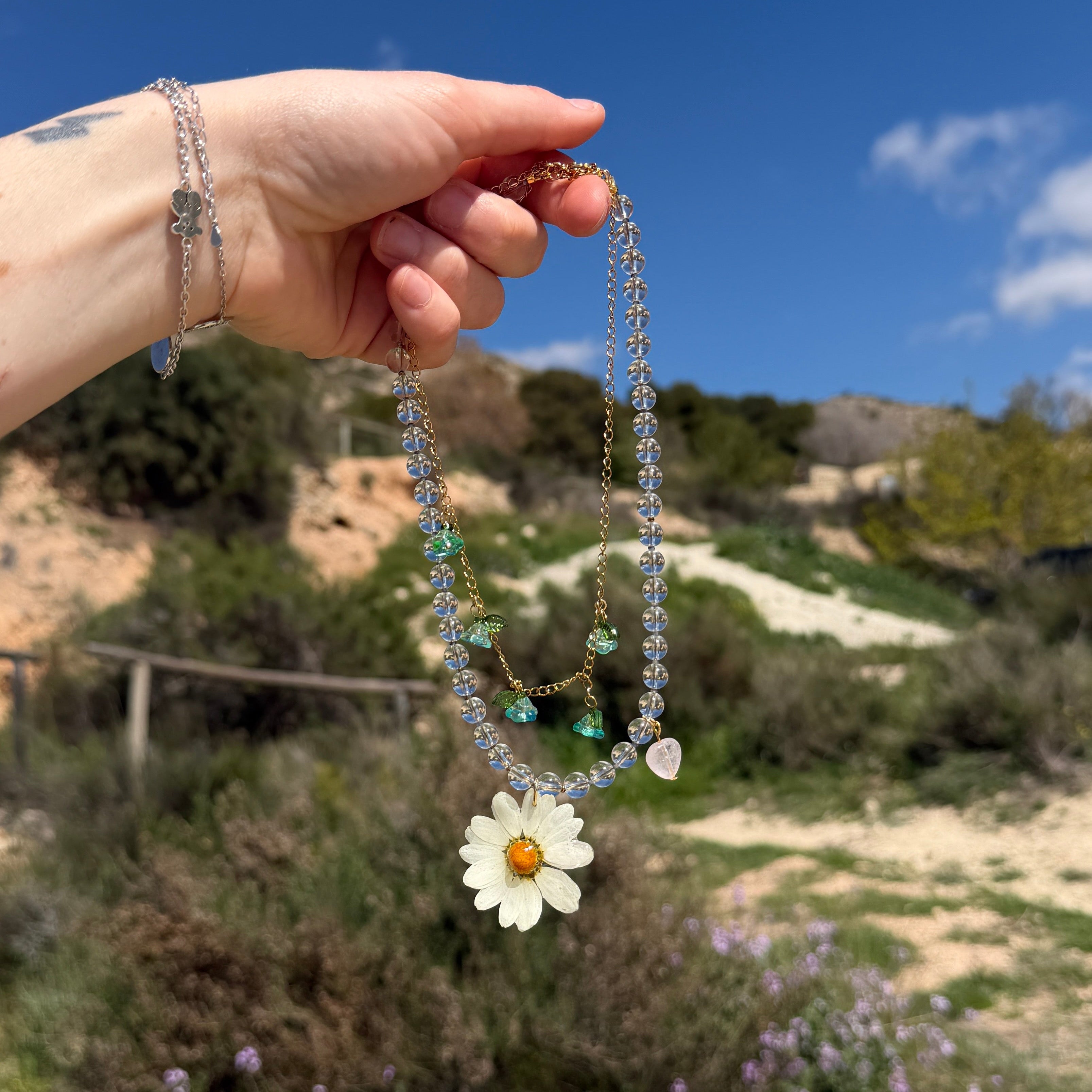Set Margarita Brisa en busto expositor entre flores silvestres amarillas, mostrando la cadena doble dorada con piedras transparentes, flores de acrílico turquesa y margarita preservada en resina