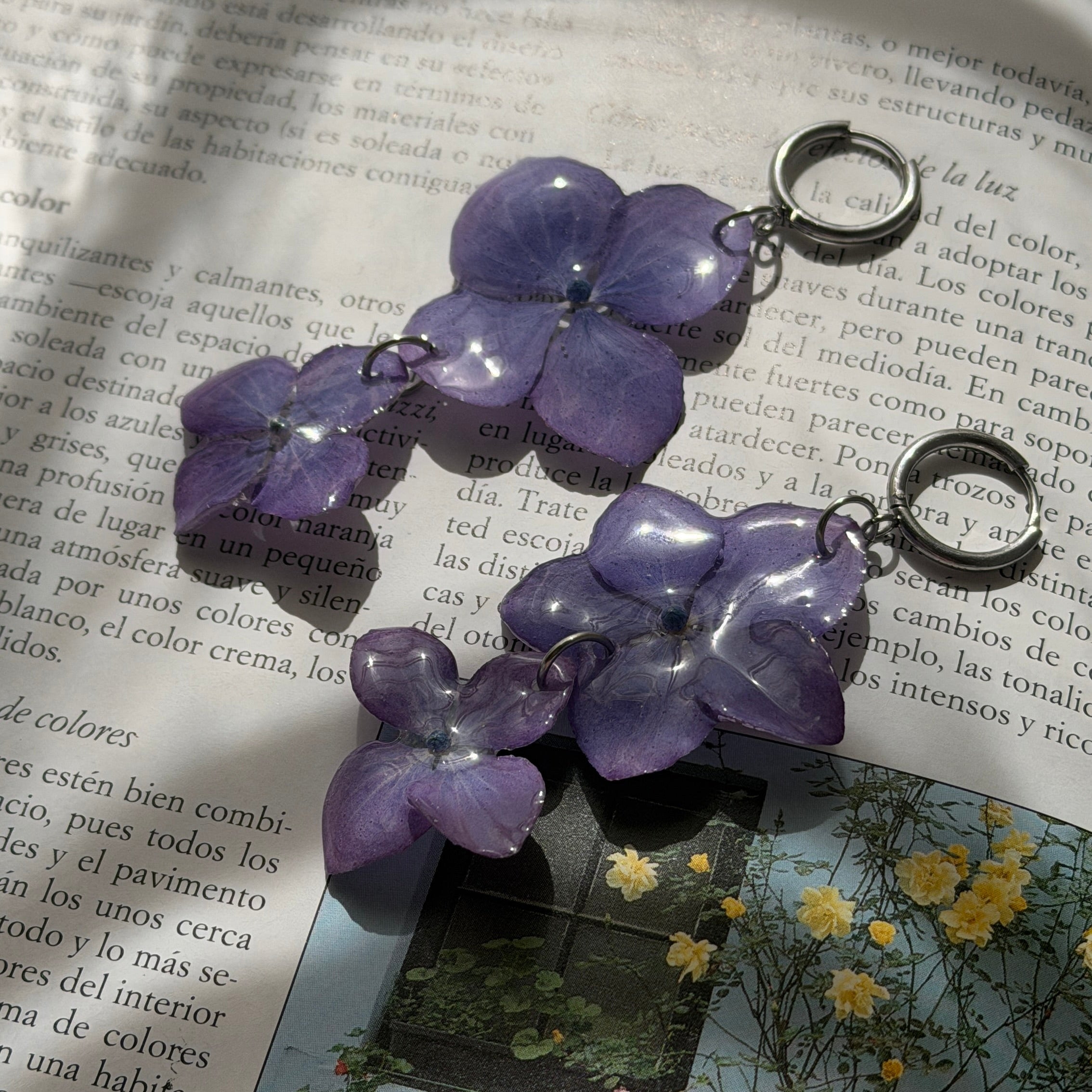Par de Pendientes Hortensia Dúo con dos flores lavanda apiladas, aros oscuros, sobre libro abierto