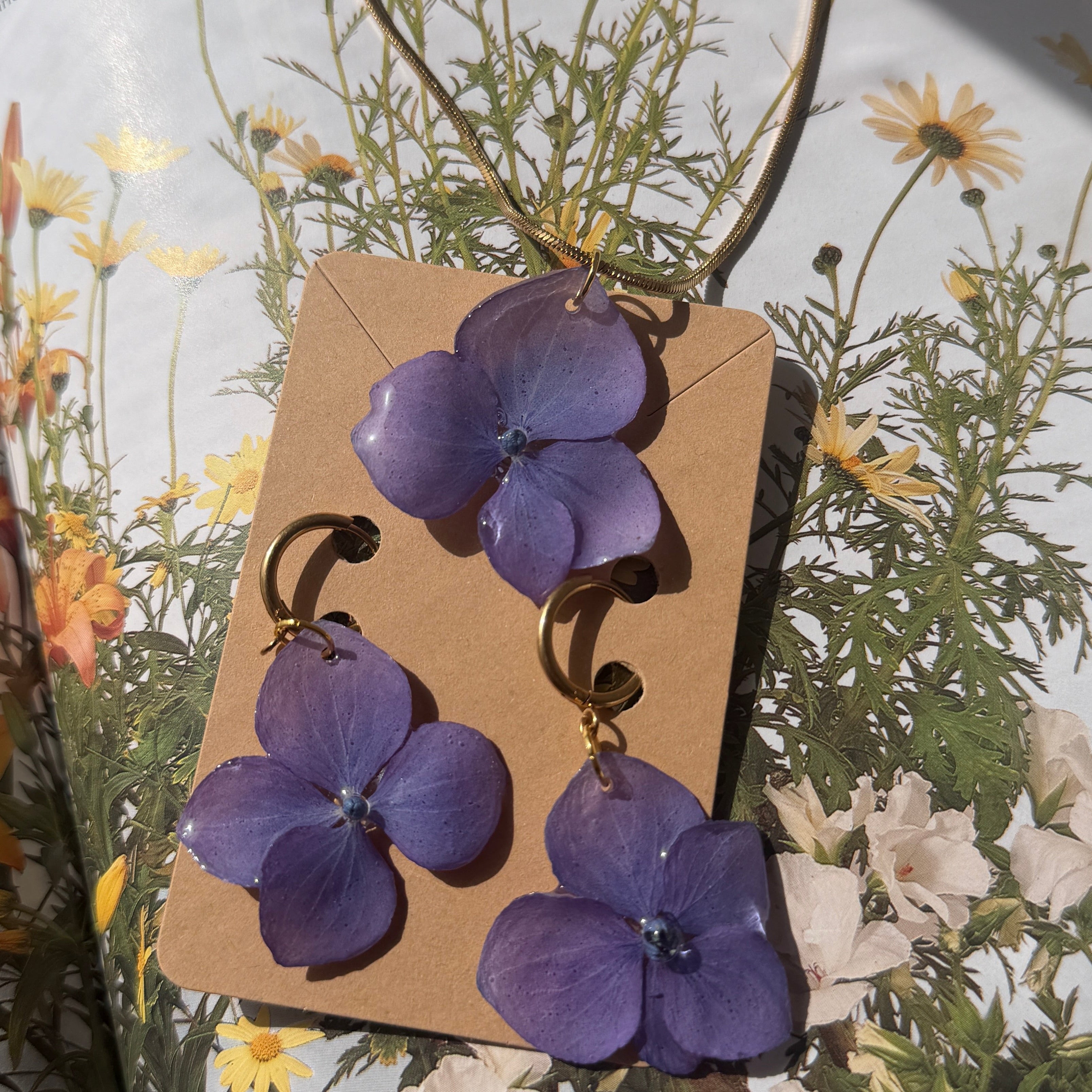 Conjunto collar y pendientes de hortensia lavanda en resina sobre tarjeta kraft con fondo floral