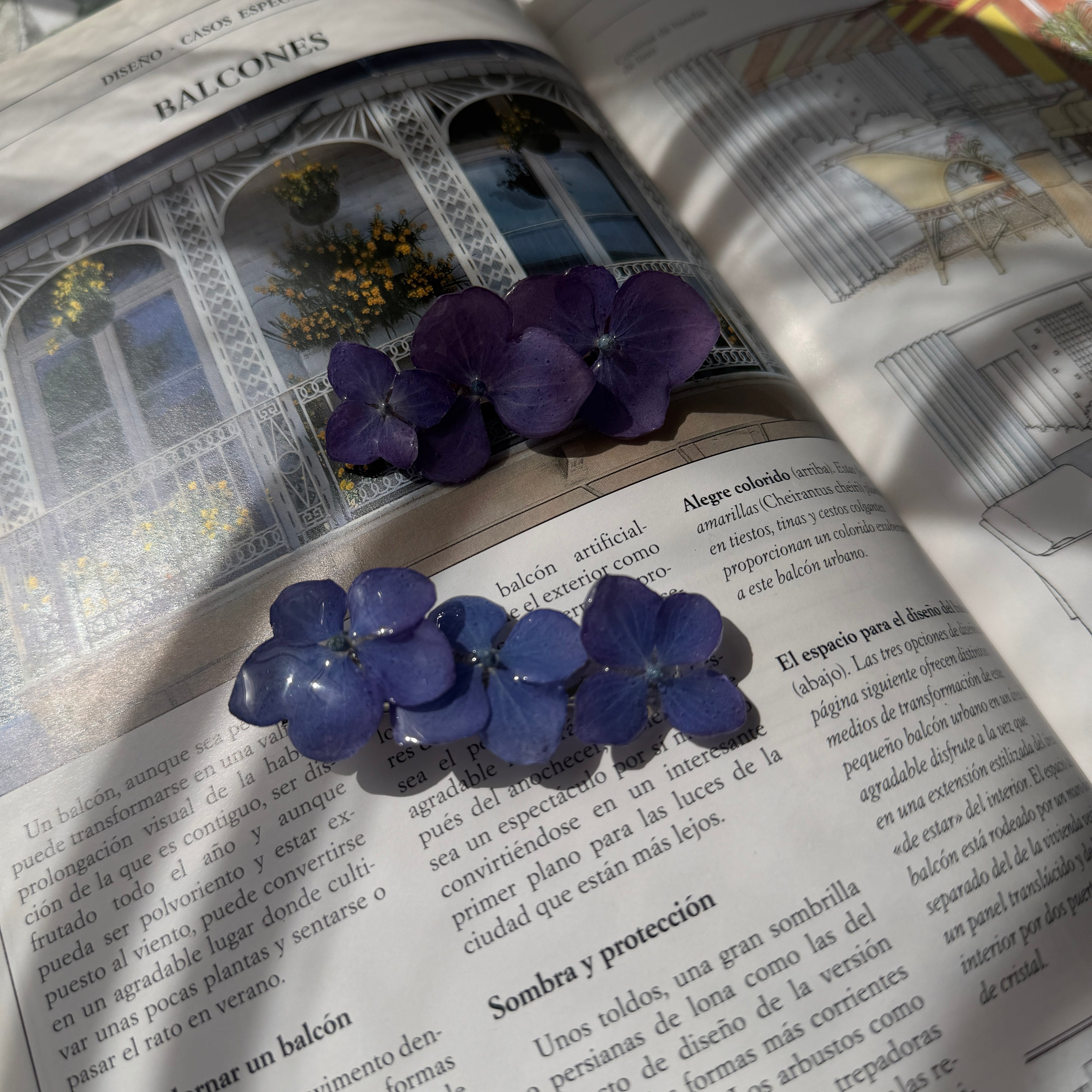 Dos pinzas de pelo con hortensias lavanda y azul sobre libro abierto con jardín, vista conjunto