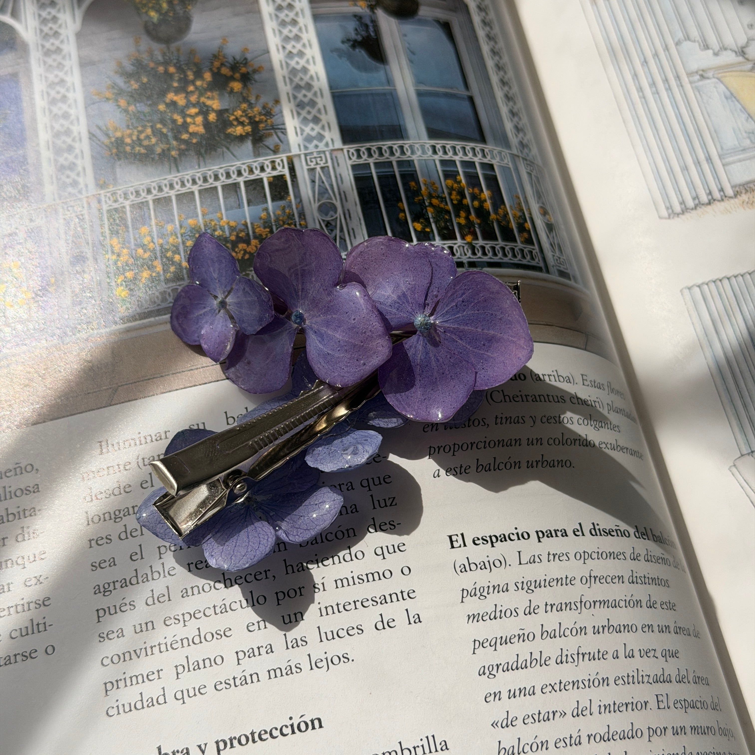 Pinza de pelo con dos flores de hortensia lavanda violeta sobre libro abierto con foto de balcón