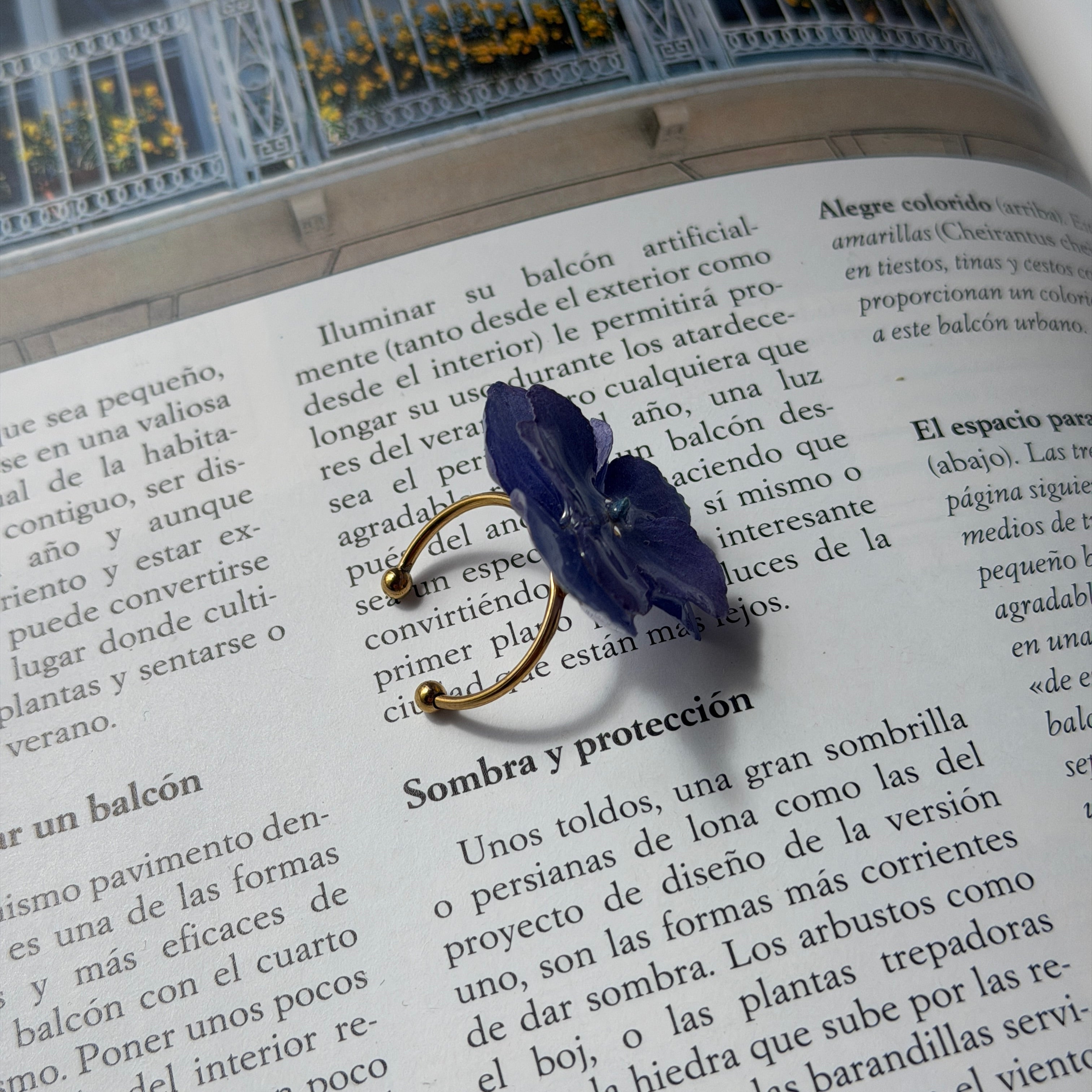 Anillo de hortensia lavanda en resina con aro abierto dorado posado sobre libro abierto, vista lateral