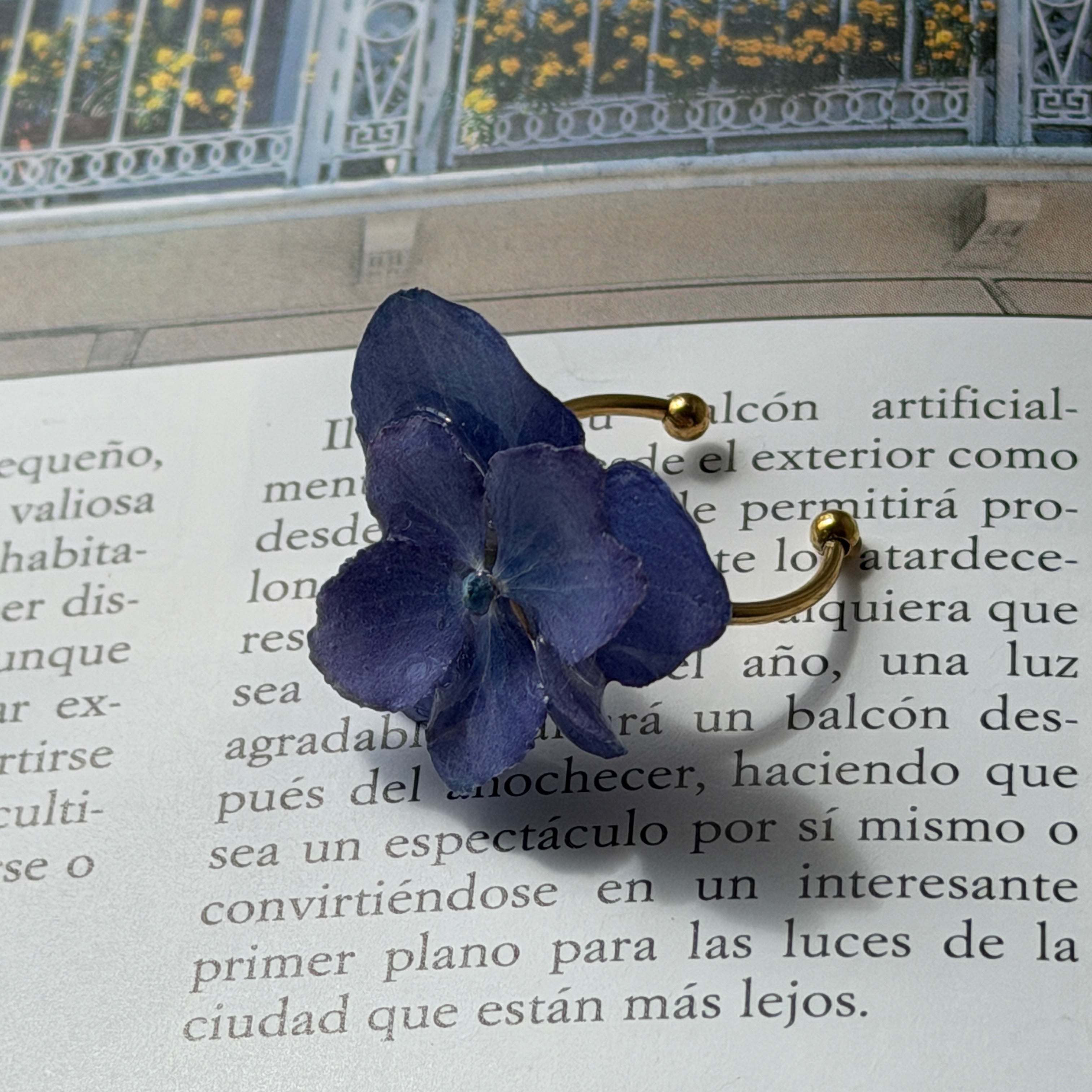 Anillo de hortensia lavanda en resina con aro dorado posado sobre libro abierto con foto de balcón