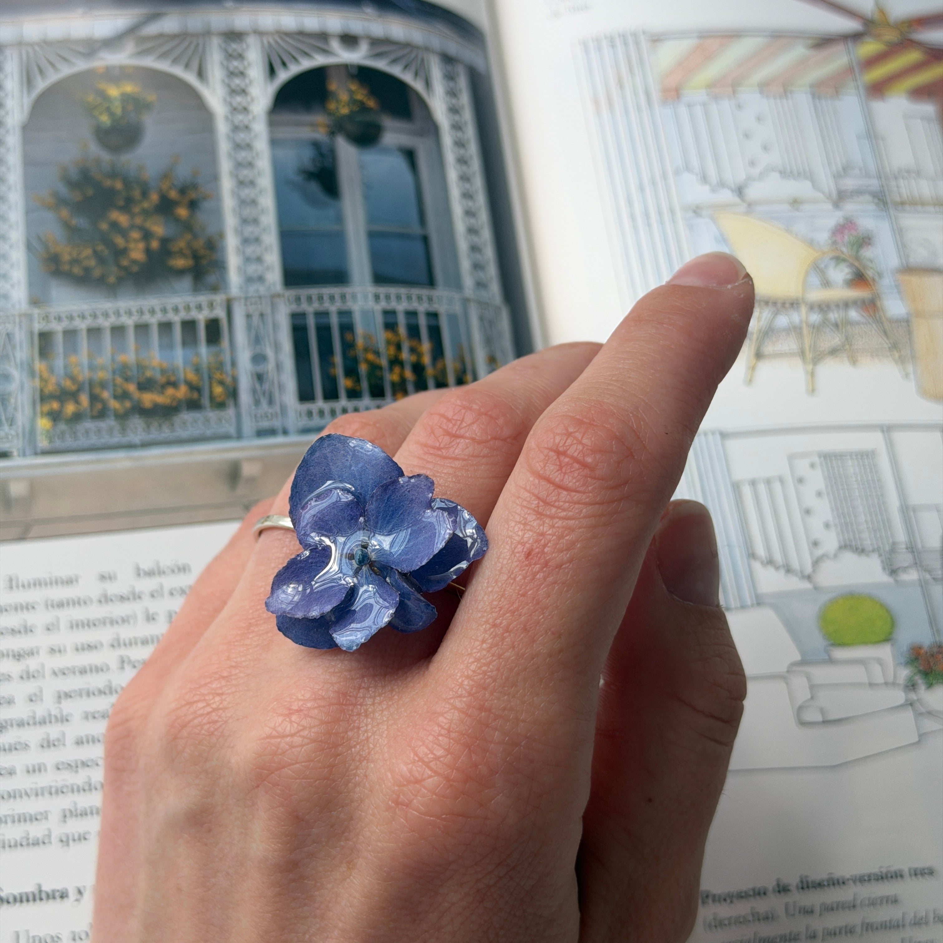 Mano de modelo con anillo de hortensia azul lavanda en resina sobre libro abierto con jardín al fondo