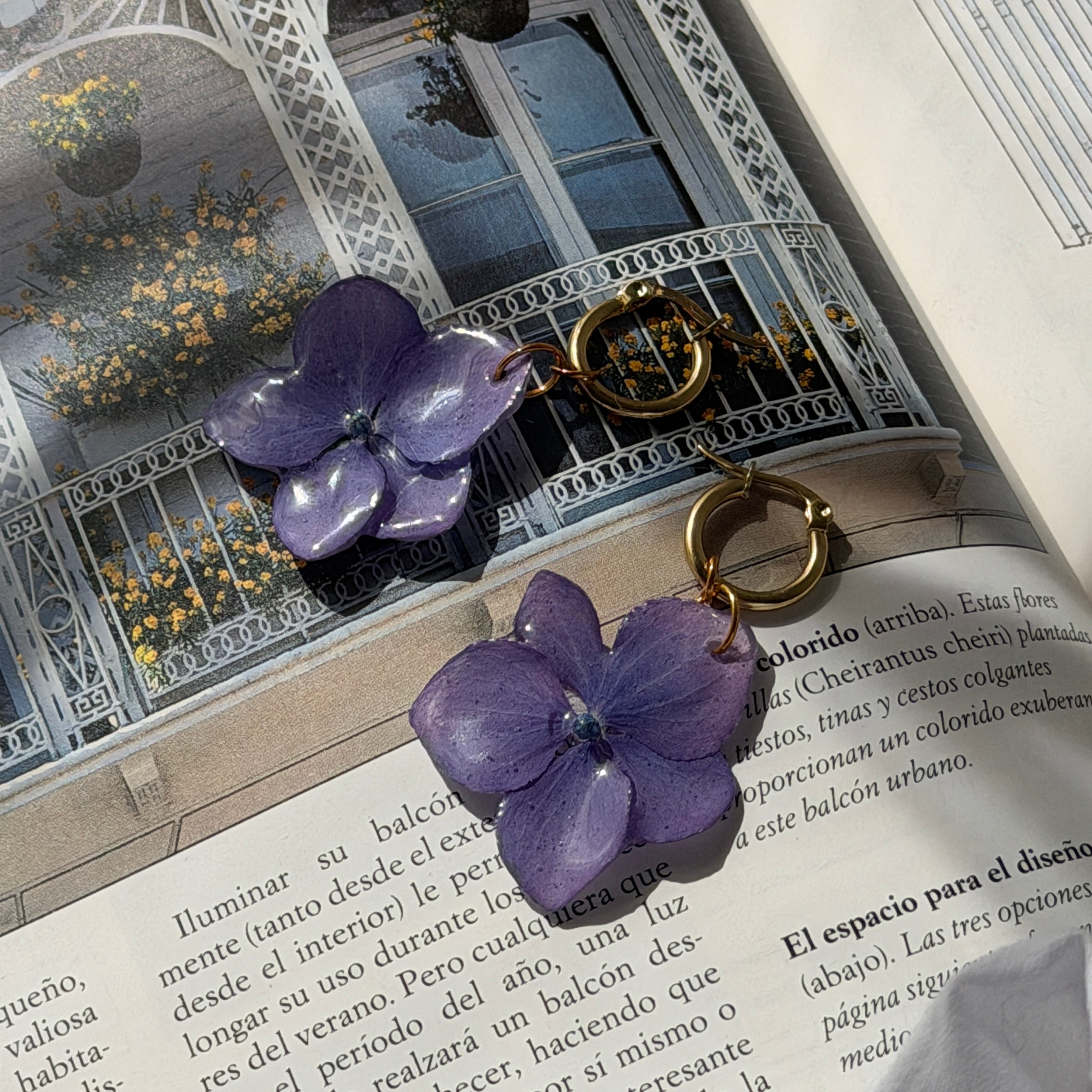 Par de pendientes de hortensia lavanda en resina con aros dorados sobre libro abierto con balcón
