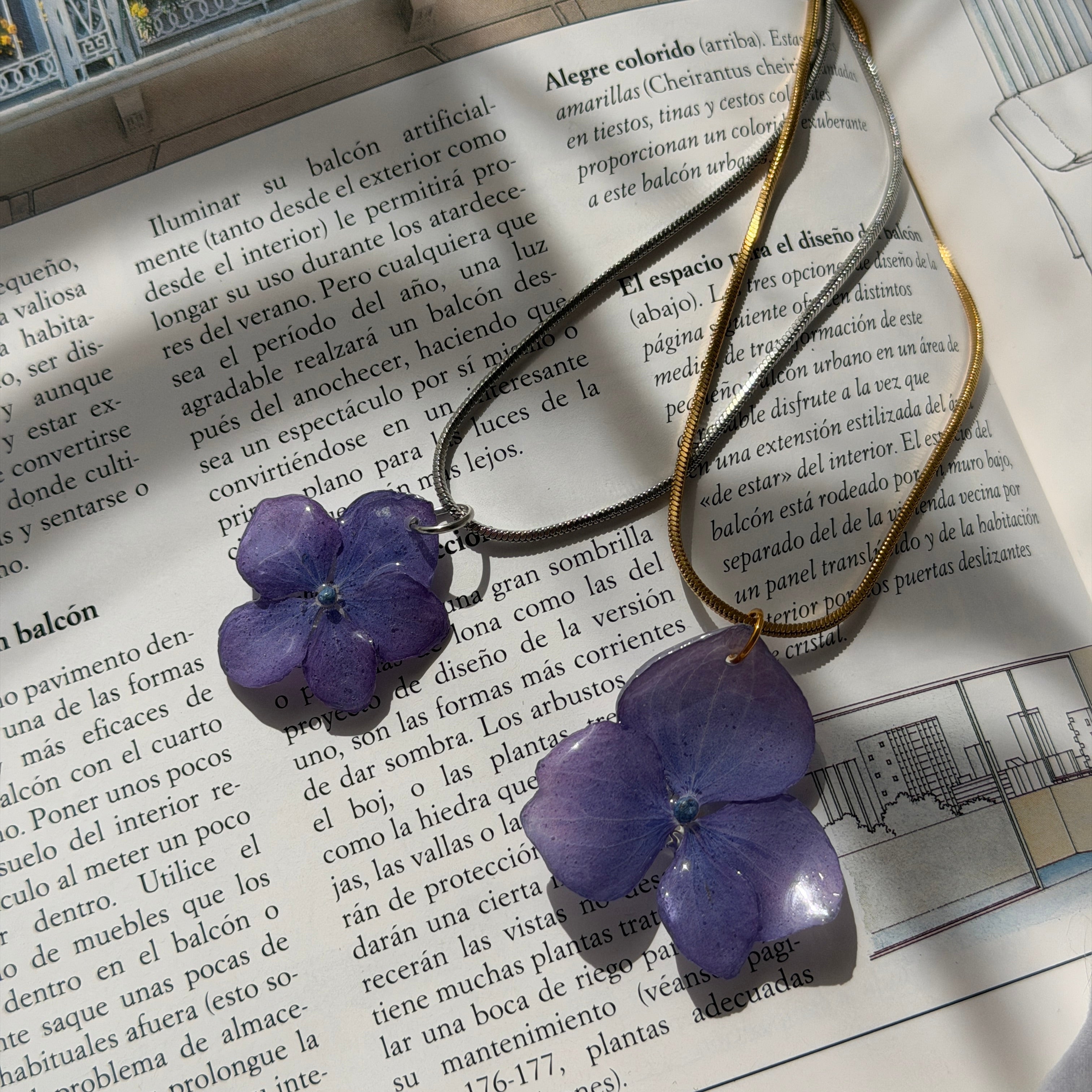 Collar Hortensia Lavanda con colgante azul violeta en resina y cadena dorada sobre libro abierto