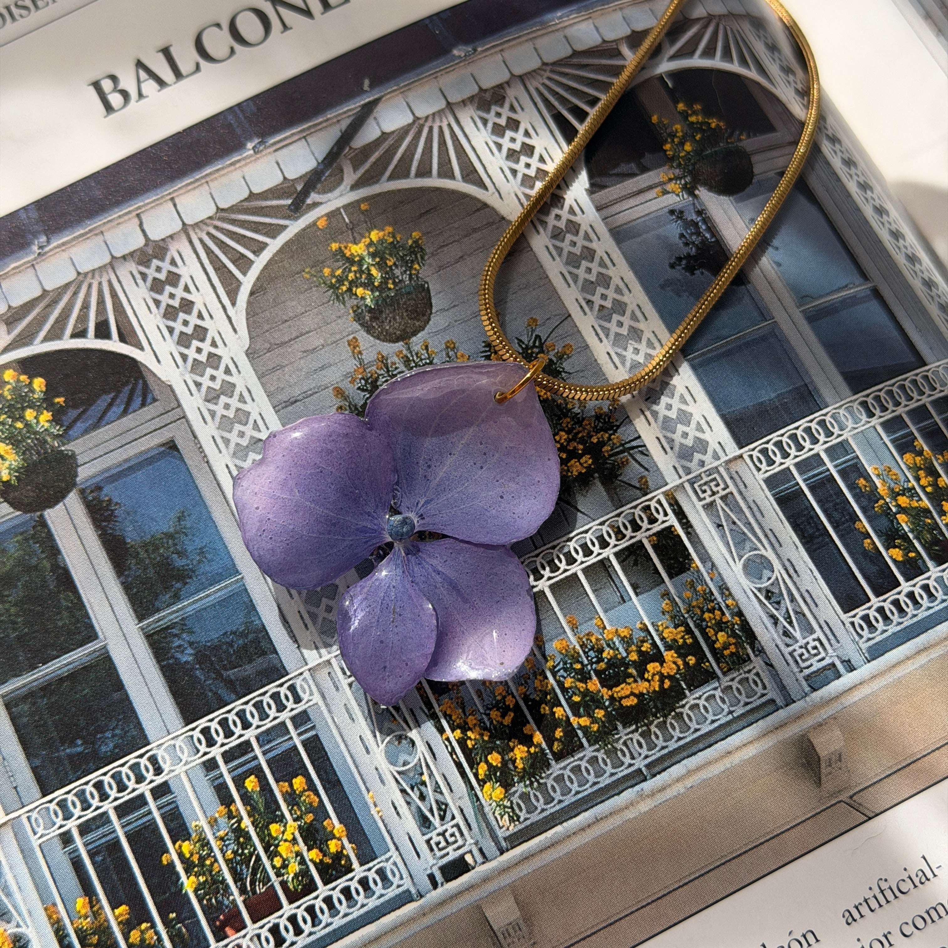 Collar de hortensia lavanda en resina con cadena dorada posado sobre libro abierto con balcón al fondo