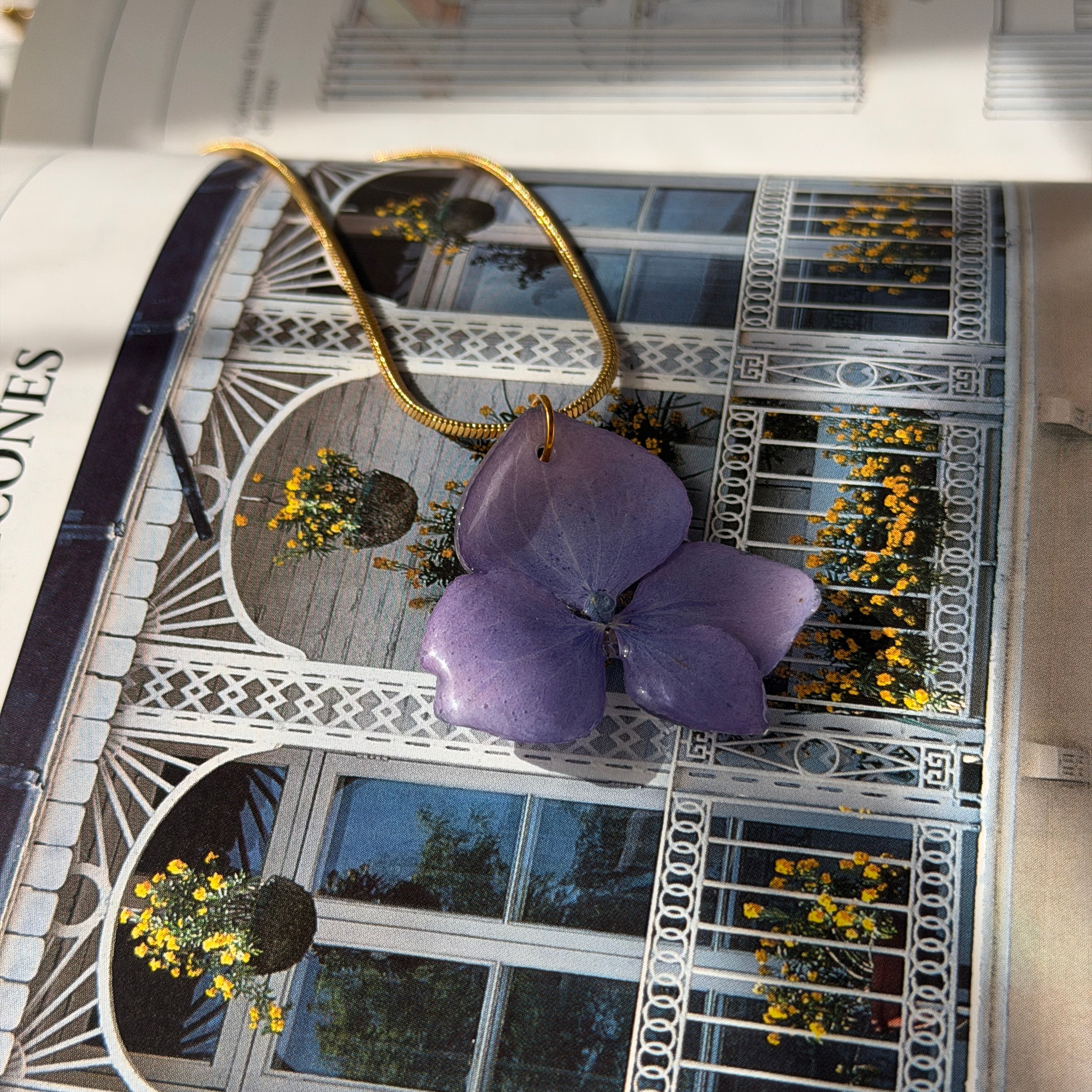 Dos variantes del Collar Hortensia Lavanda, cadena plateada y dorada, posadas sobre libro abierto