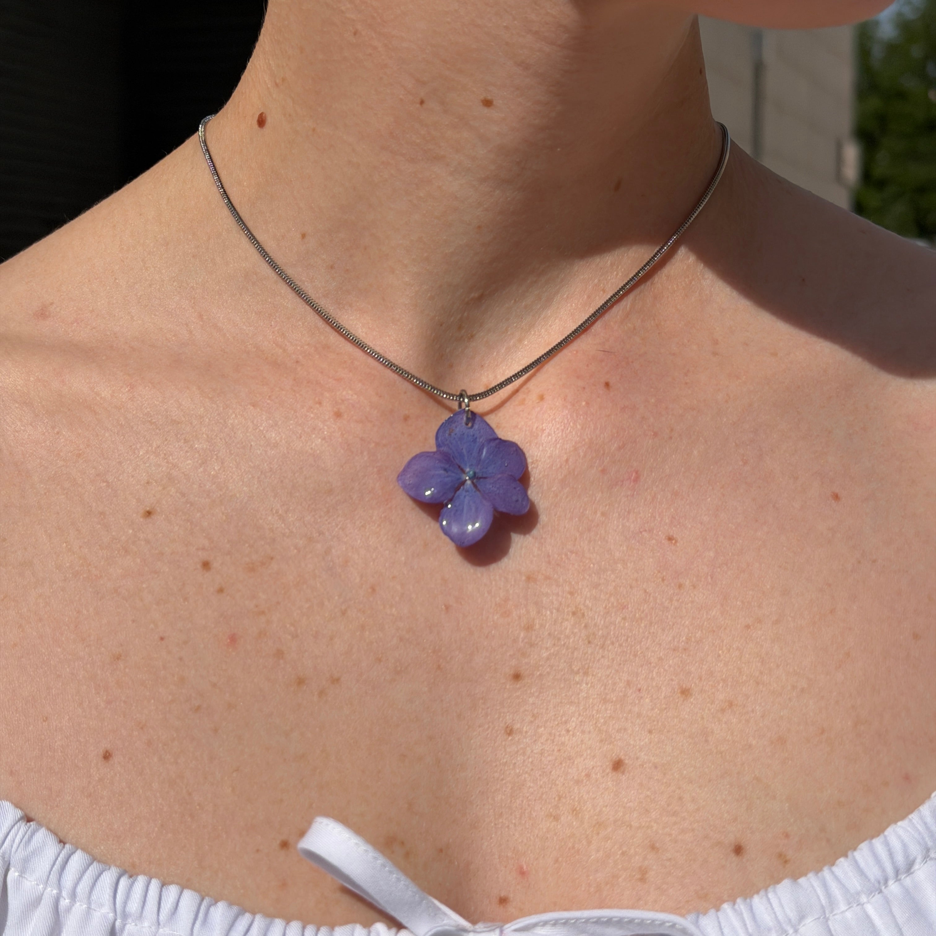 Modelo luciendo collar de hortensia lavanda en resina con cadena plateada al sol, vista del escote