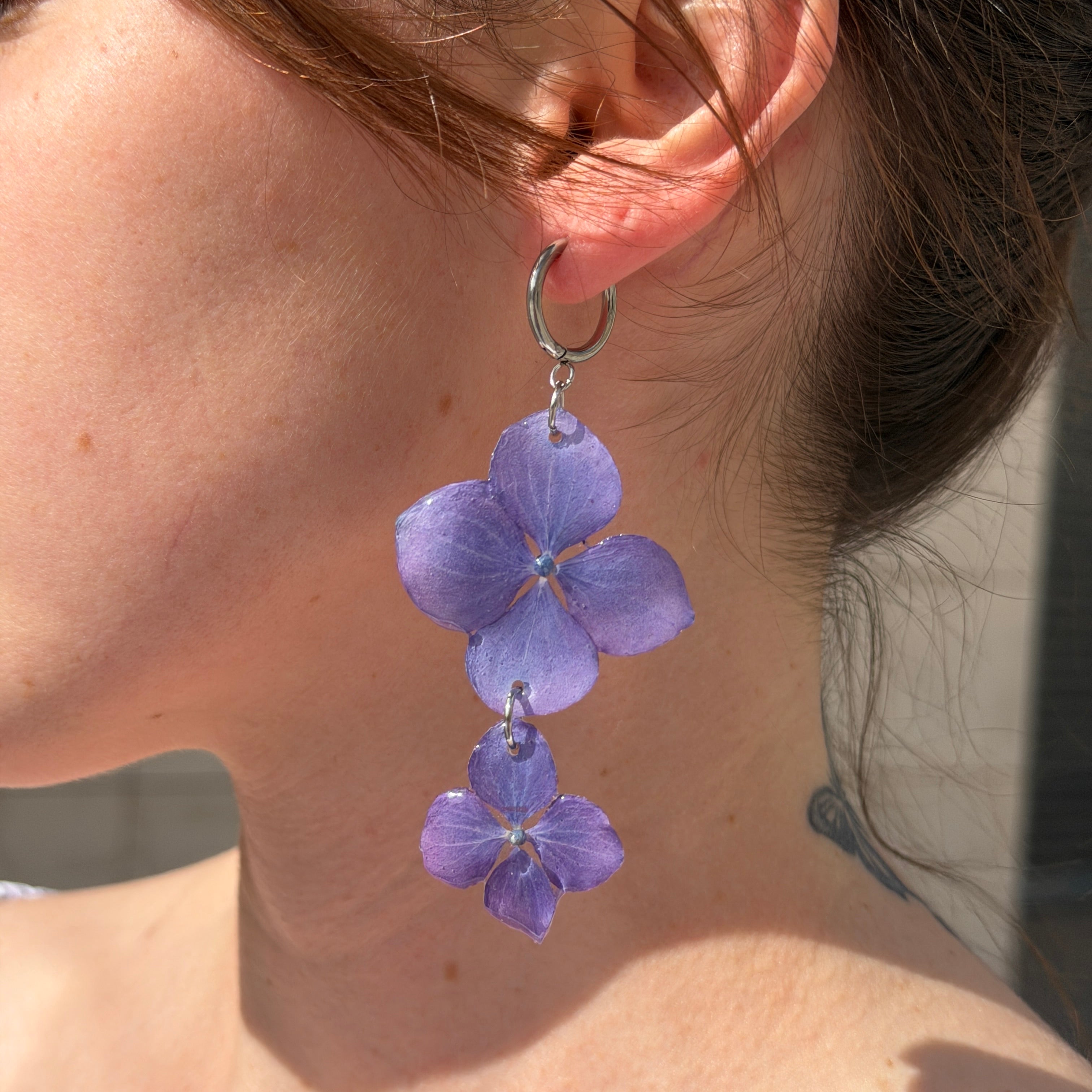 Modelo con Pendientes Hortensia Dúo en oreja, dos hortensias lavanda apiladas en aro plateado al sol