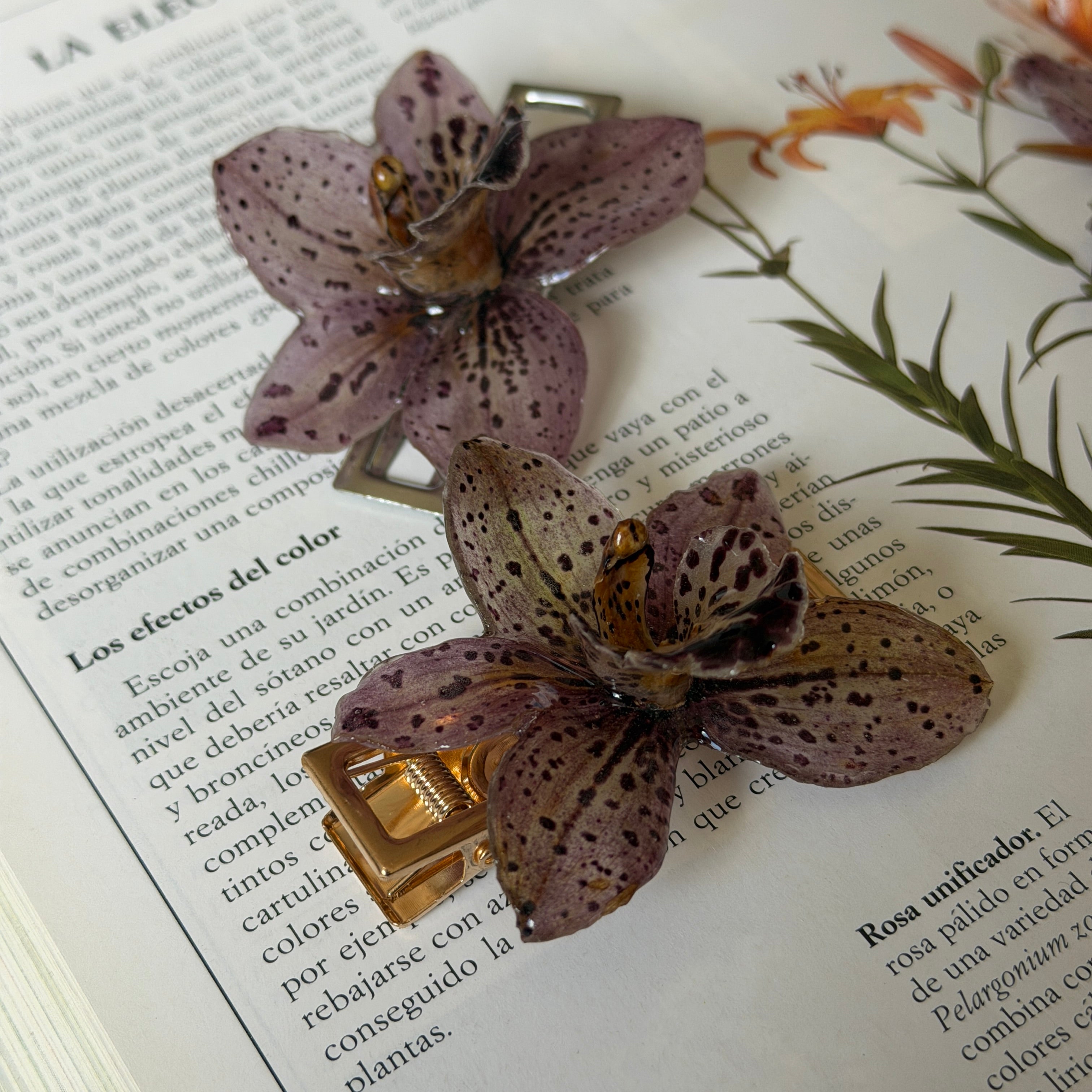 Dos pinzas de orquídea Velaria, una dorada y una plateada, sobre libro de jardín con flores naranjas