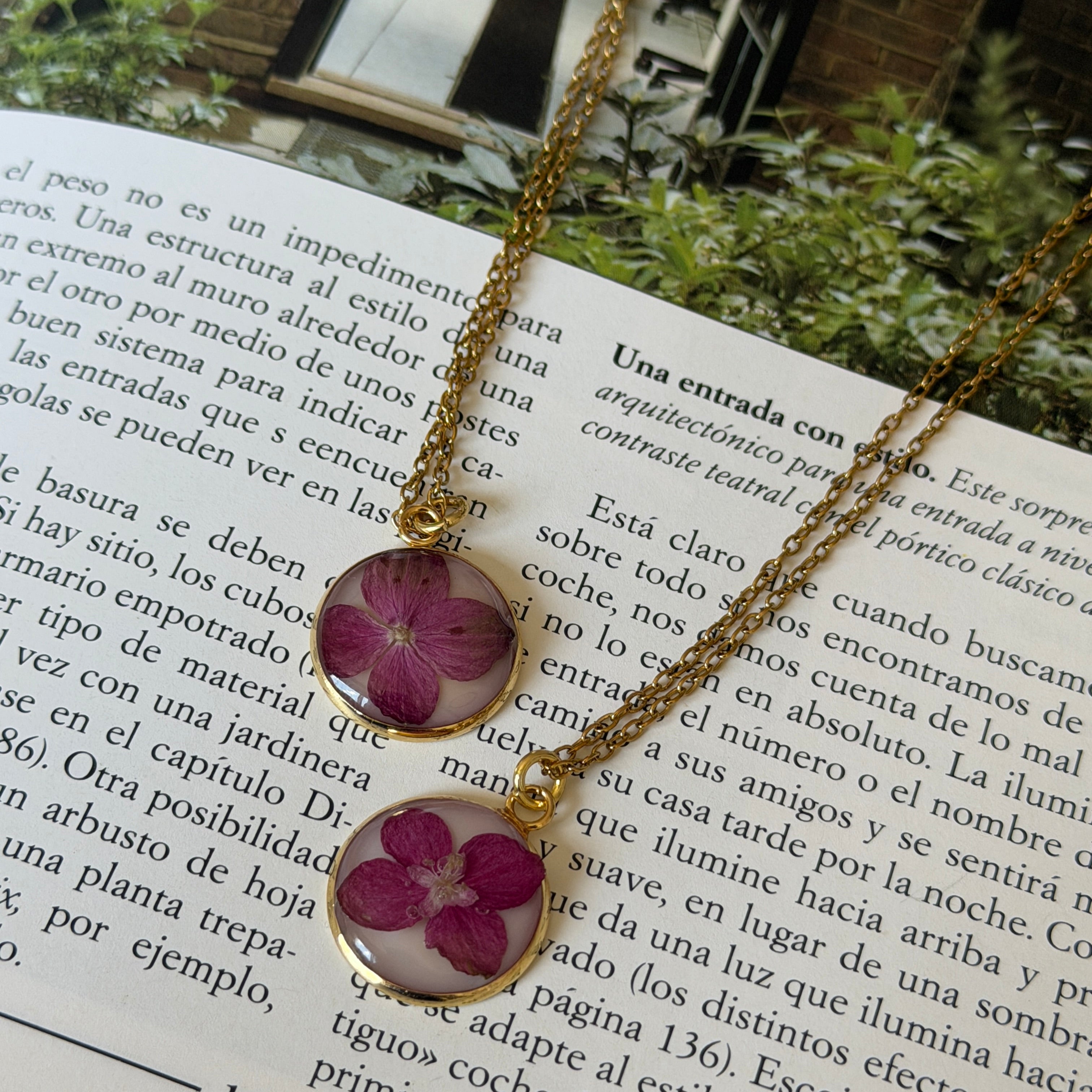 Dos collares Hortensia Mireia sobre libro con jardín, medallones dorados circulares con hortensia fucsia