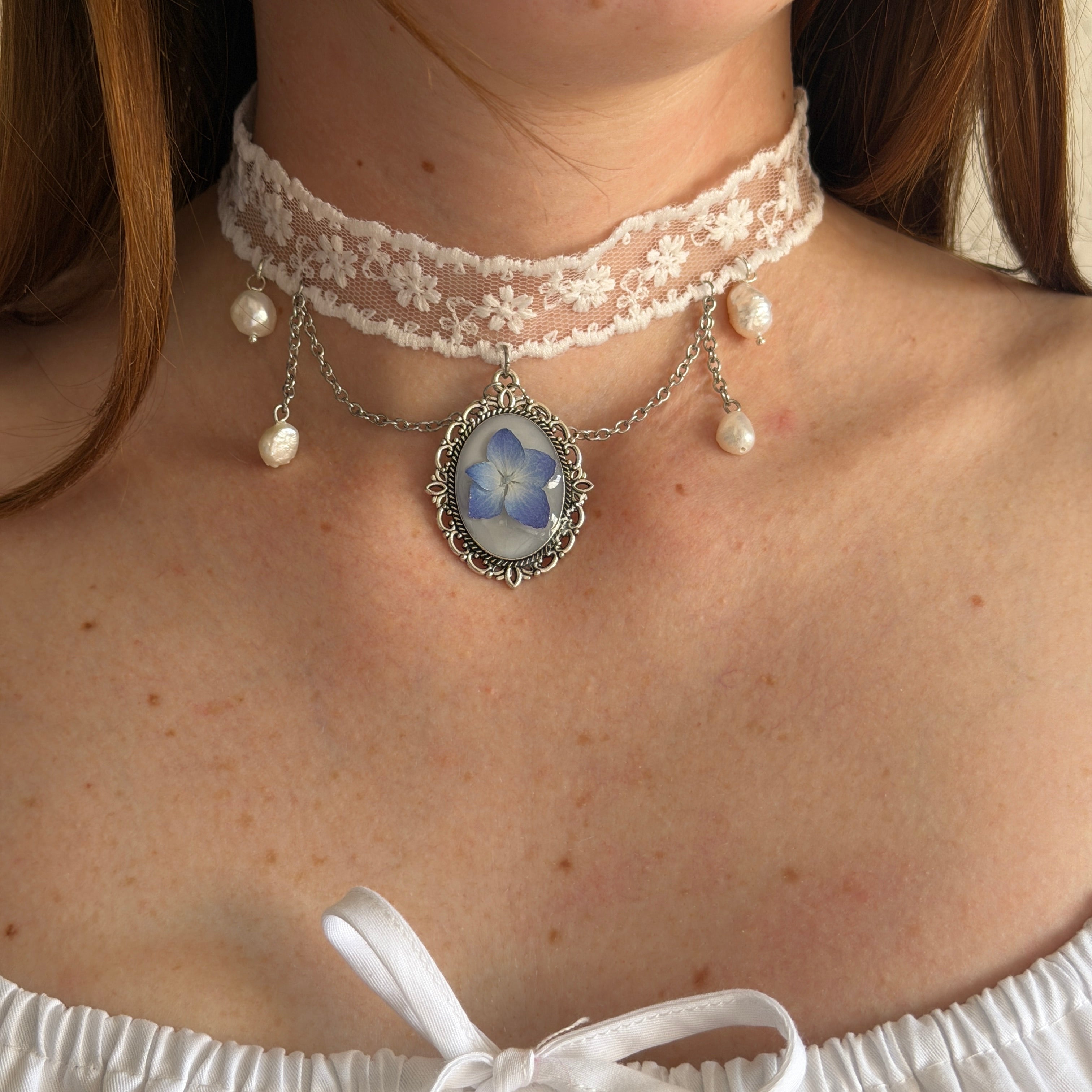Modelo con Gargantilla Leticia: encaje blanco con medallón oval plateado, hortensia azul y perlas