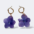 Par de pendientes de hortensia lavanda preservada en resina colgando de aros dorados, fondo gris