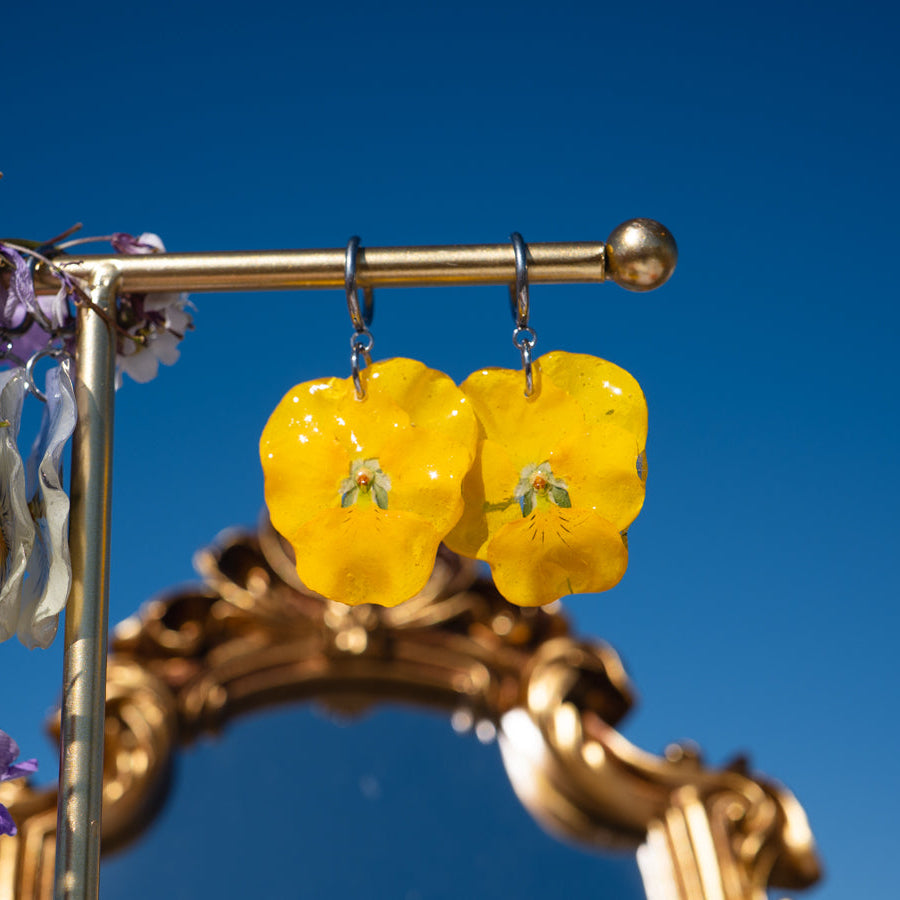 Par de Pendientes Pensamiento Candela amarillo colgando de barra frente a espejo dorado vintage, cielo azul