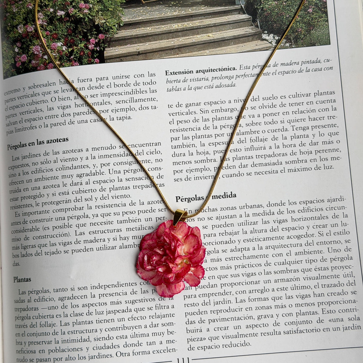 Collar Clavel Júpiter, clavel fucsia encapsulado en resina, sobre un libro de jardinería con fotografía de pérgola