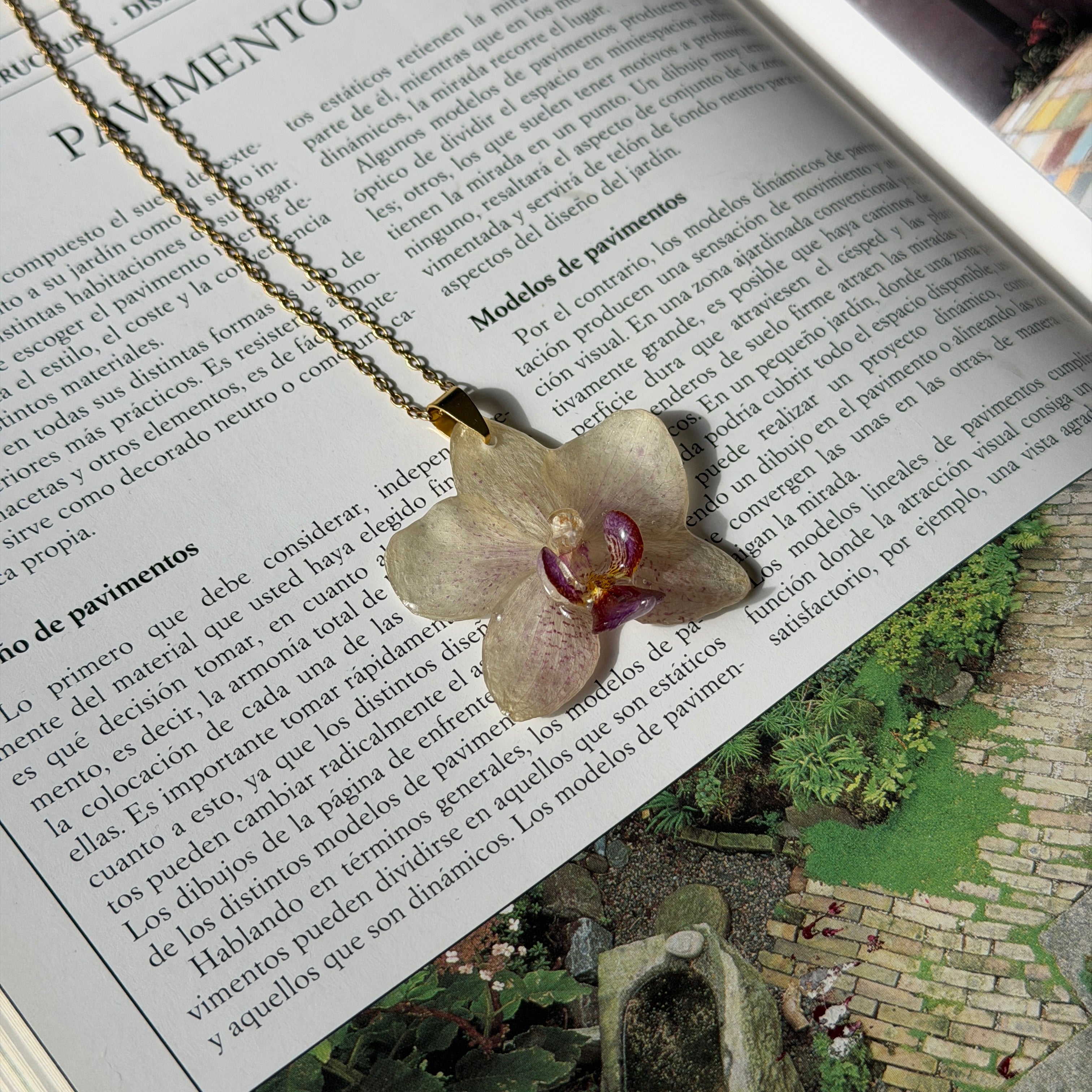 Collar con orquídea blanco marfil encapsulada en resina, sobre cadena de acero inoxidable, apoyado sobre revista de jardinería