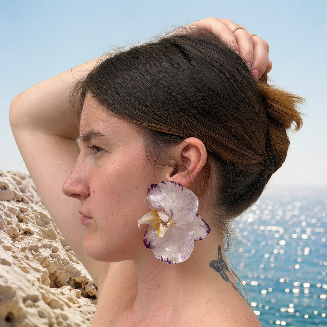 Mujer llevando pendientes de orquídea blanca y violeta encapsulada en resina sobre la oreja, junto al mar