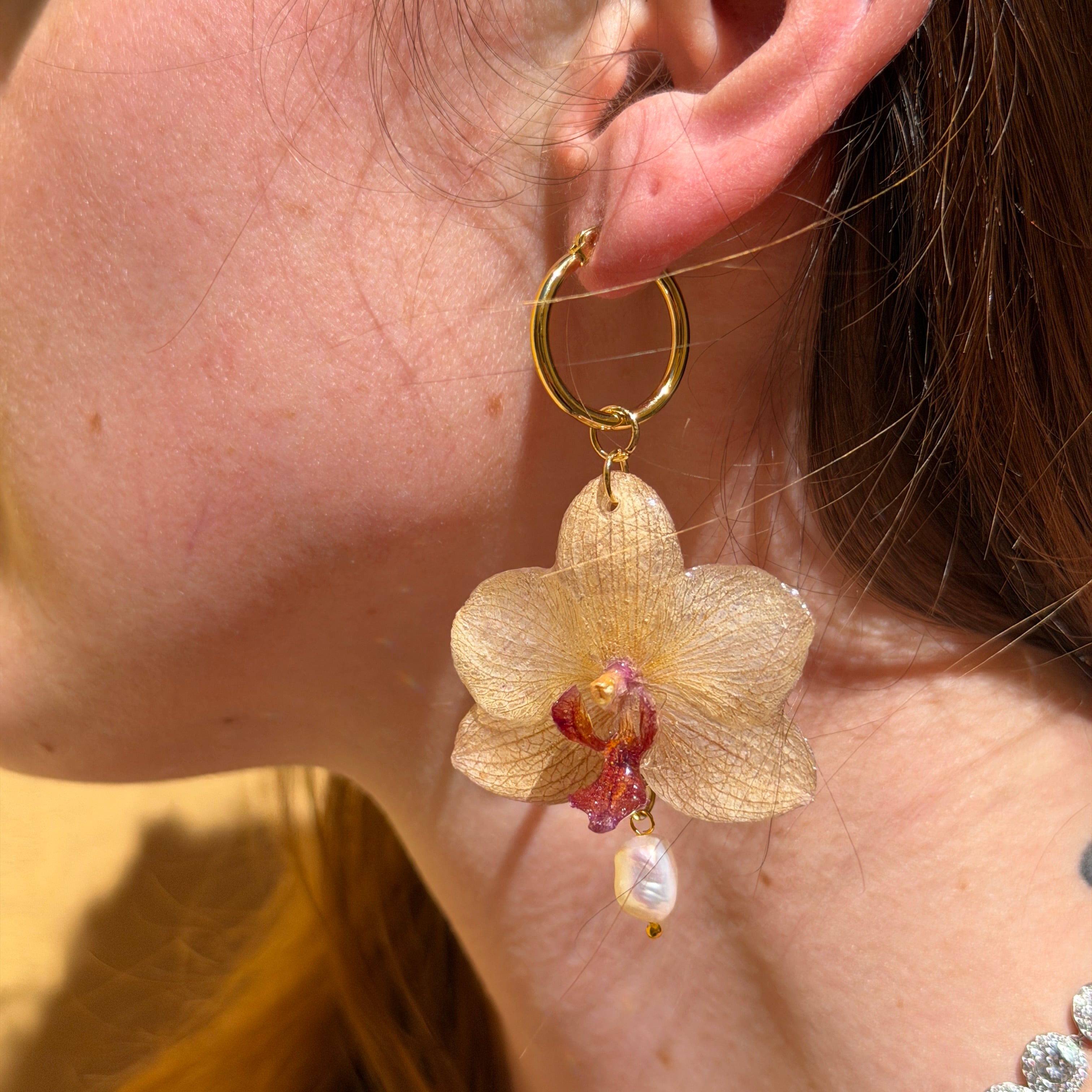 Mujer llevando pendiente de orquídea marfil tostado encapsulada en resina sobre la oreja, con perla natural colgante