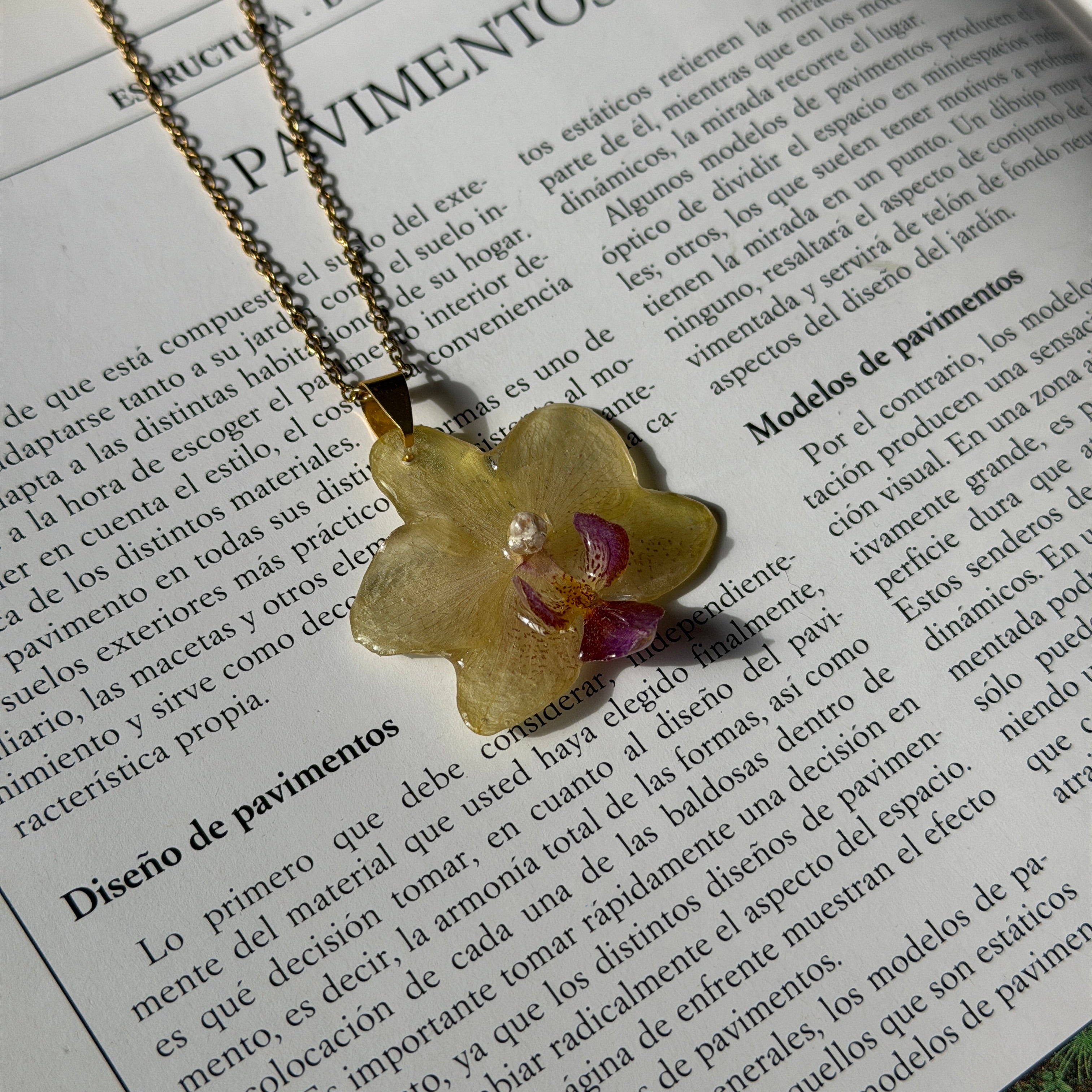 Collar con orquídea amarillo mantequilla encapsulada en resina, sobre cadena de acero inoxidable, apoyado sobre revista de jardinería