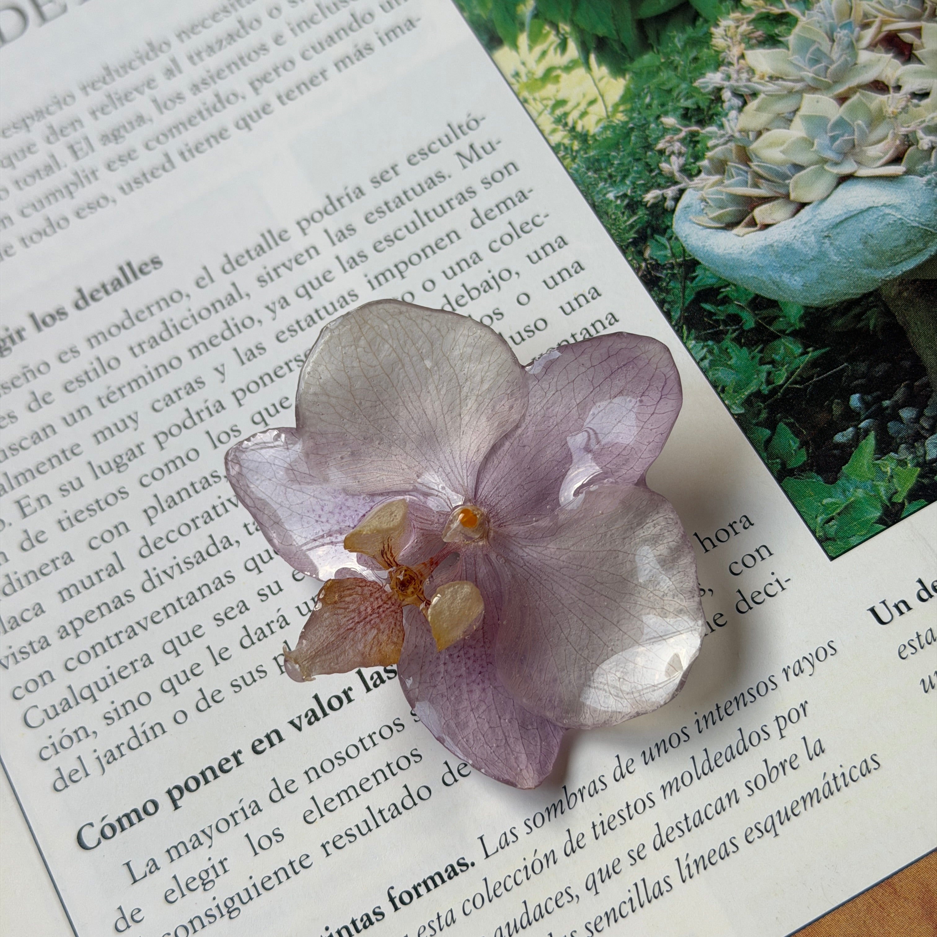 Pinza de orquídea lavanda encapsulada en resina sobre una página de revista de jardinería