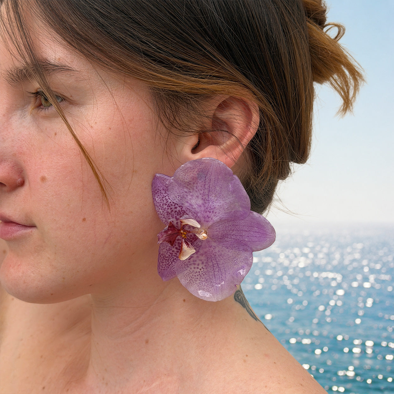 Detalle de pendiente de orquídea lila encapsulada en resina sobre la oreja, con el mar al fondo