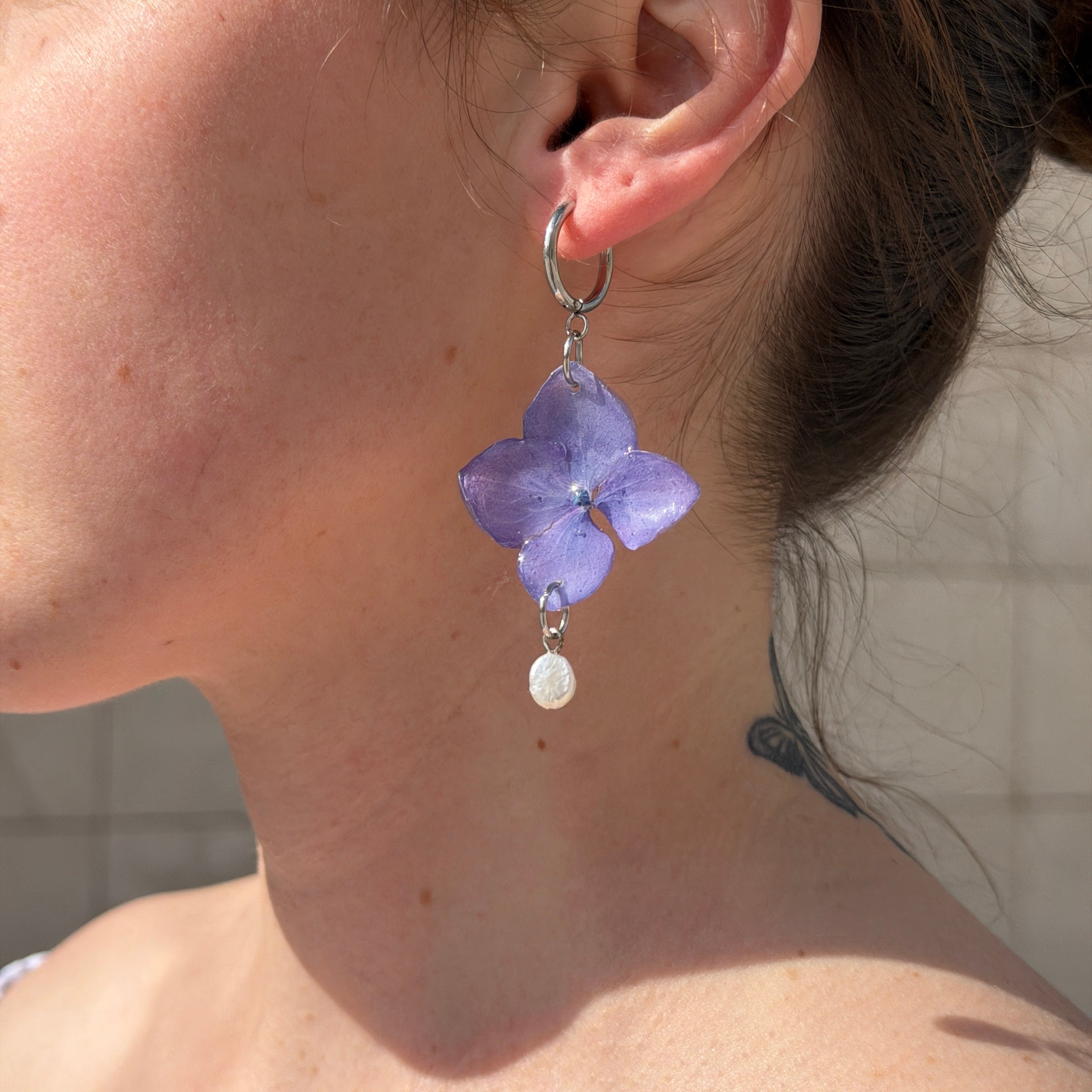 Mujer llevando pendientes de hortensia azul violáceo encapsulada en resina con perla barroca, sobre la oreja