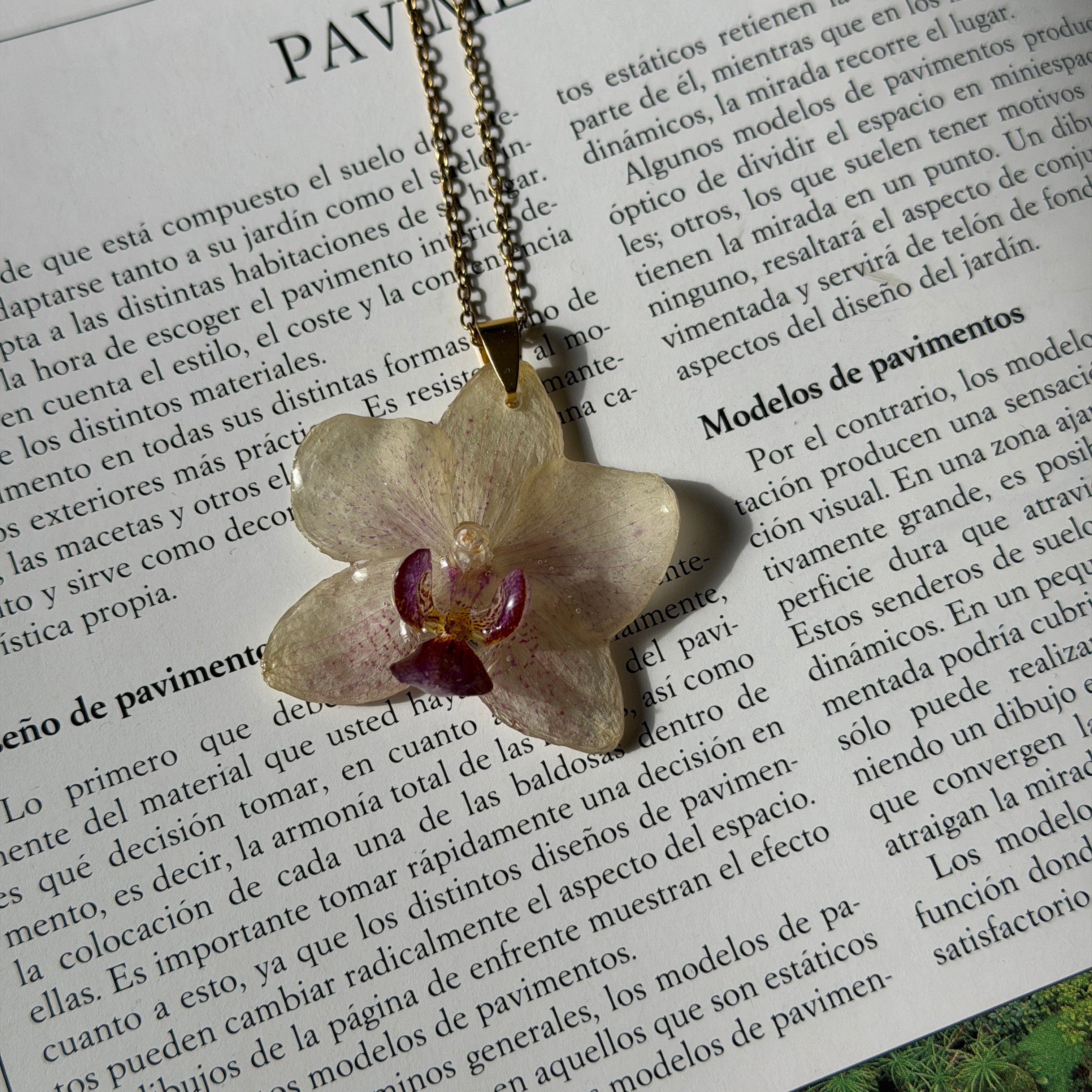 Collar con orquídea blanco marfil y labelo magenta encapsulada en resina, sobre revista abierta