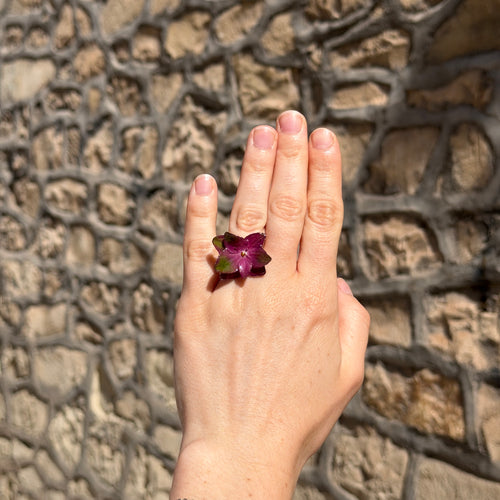 Anillo de hortensias magenta encapsuladas en resina en el dedo, sobre pared de piedra