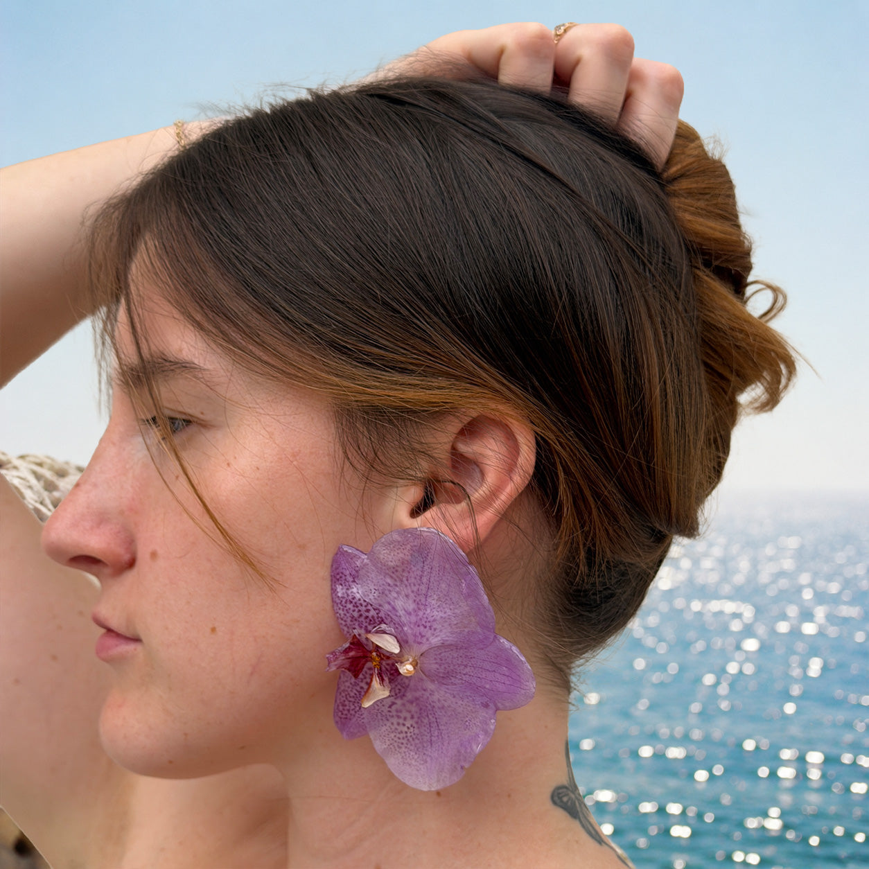 Mujer llevando pendientes de orquídea lila encapsulada en resina, de perfil junto al mar