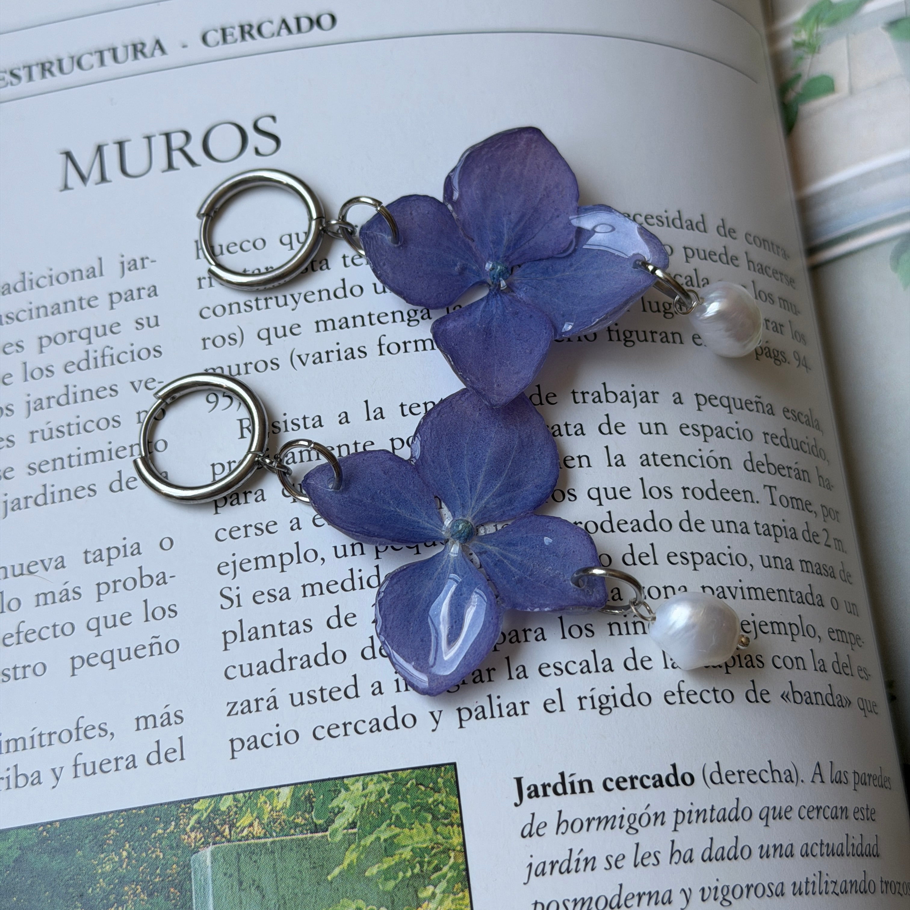 Pendientes de hortensia azul violáceo encapsulada en resina con perla barroca, sobre la página de un libro