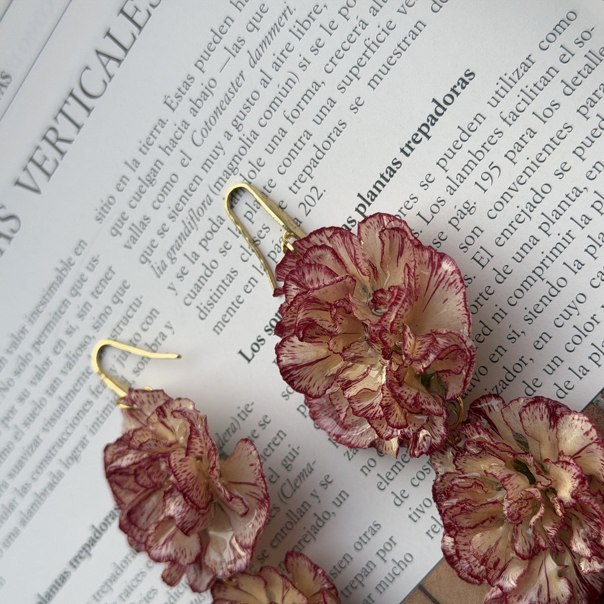 Pendientes Clavel Nácar con anzuelo dorado y dos claveles, sobre un libro de plantas